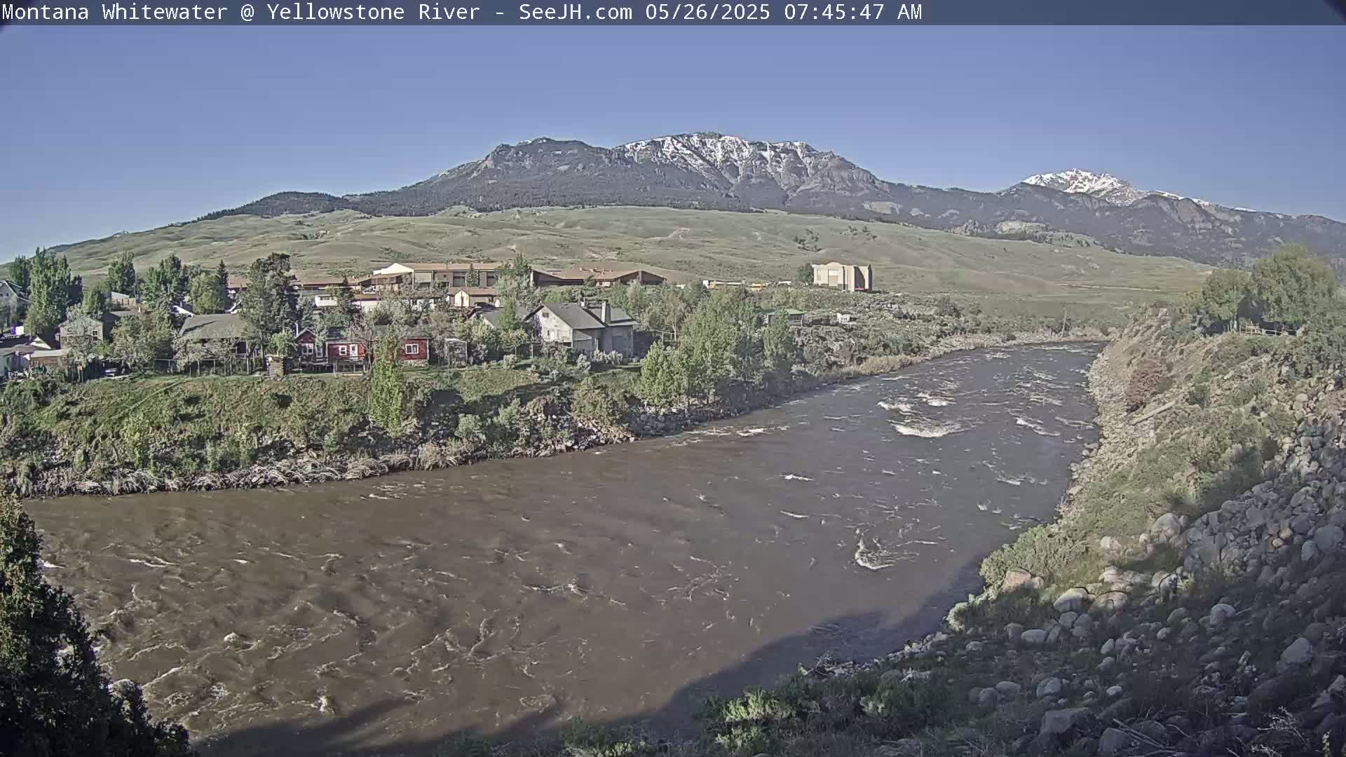 A muddy river flows past a small town nestled against snow-capped mountains under a clear sky.