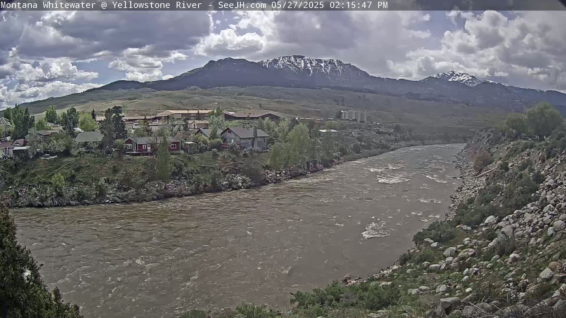 A cloudy day shows a muddy river flowing past a small town nestled at the base of snow-capped mountains.