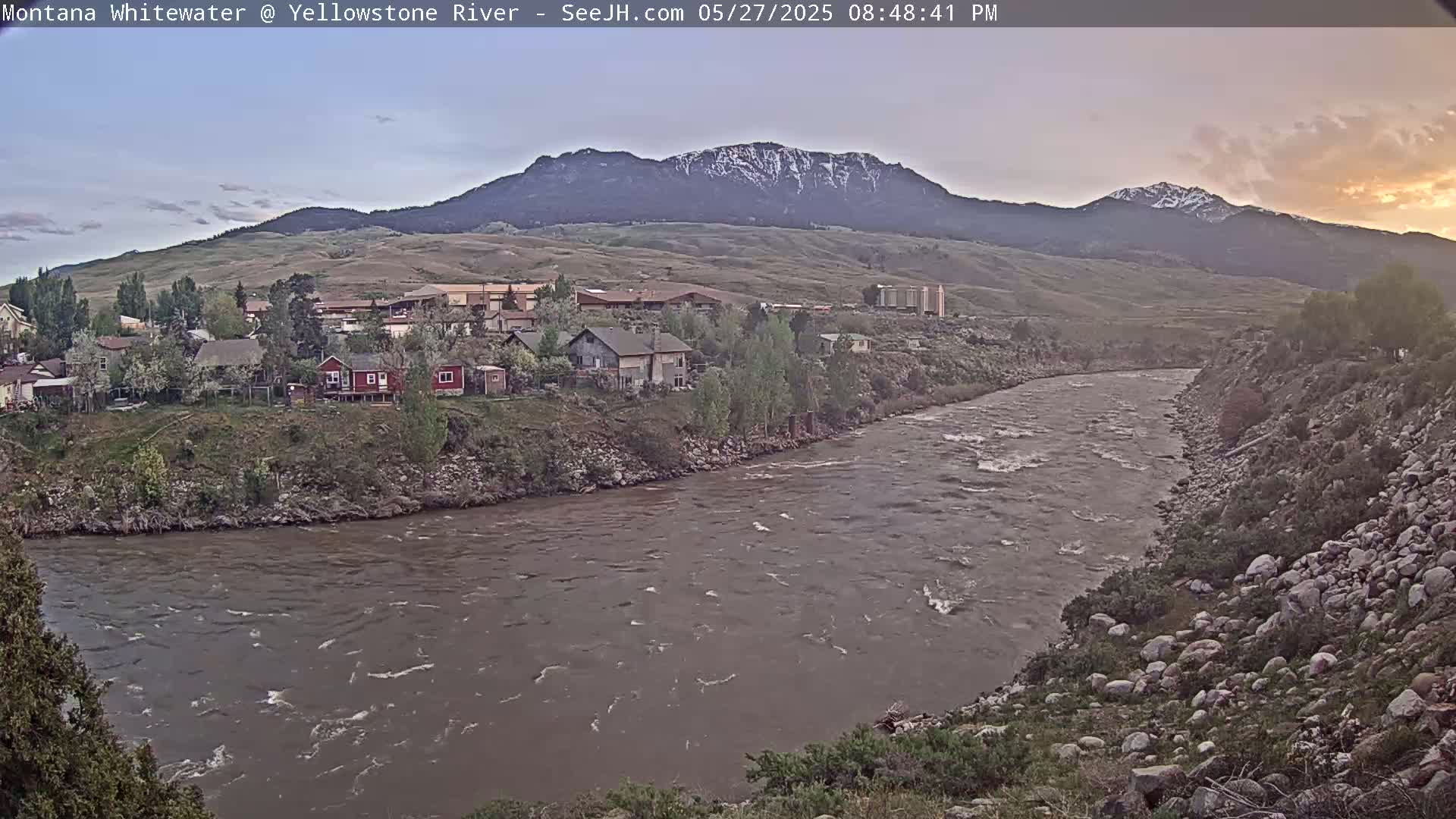 A wide, fast-flowing river curves past a small town nestled at the base of a mountain range under a partly cloudy, dusky sky.
