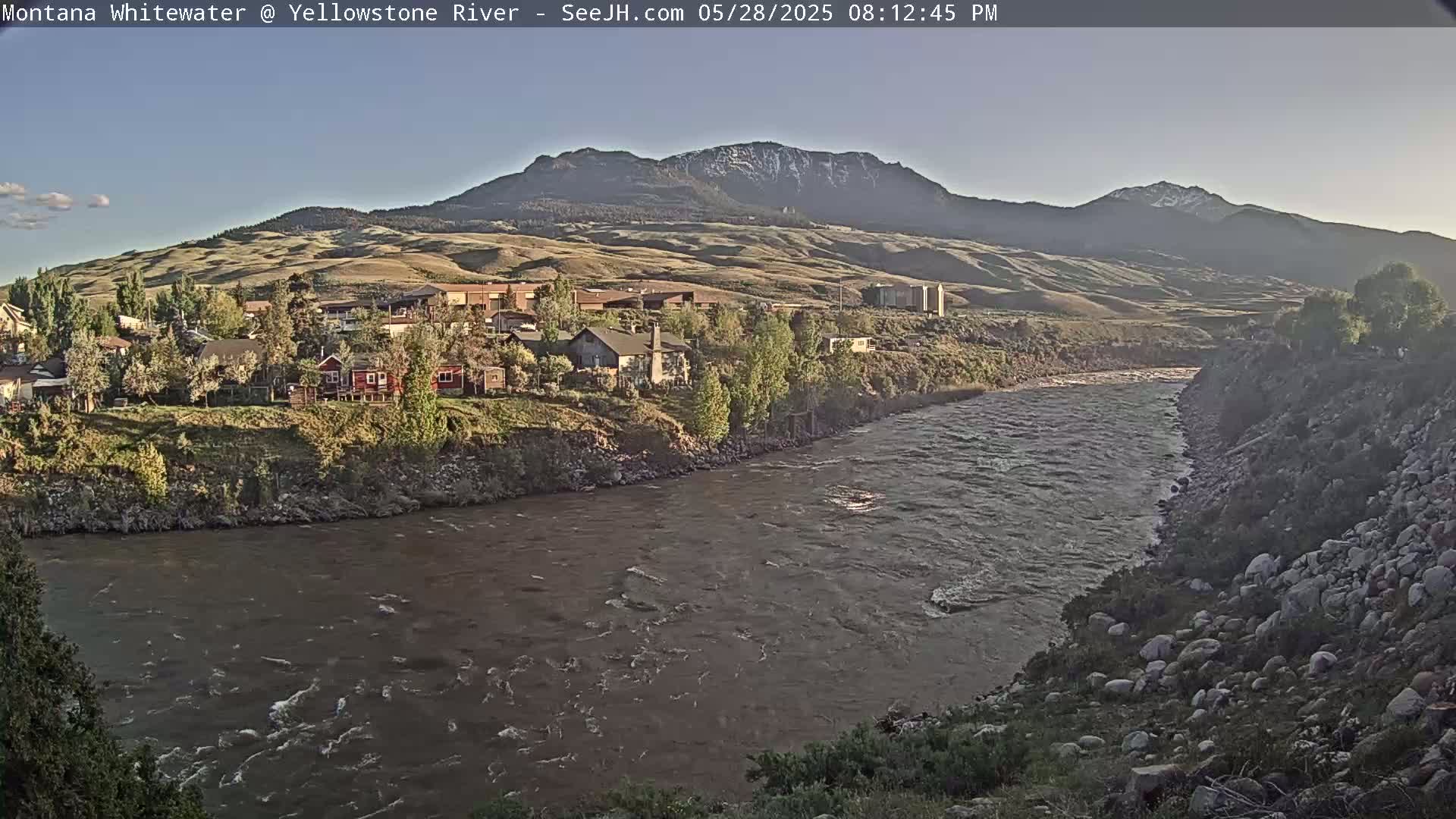 A wide, brown river flows past a small town nestled against a mountain range under a clear, light-blue sky.