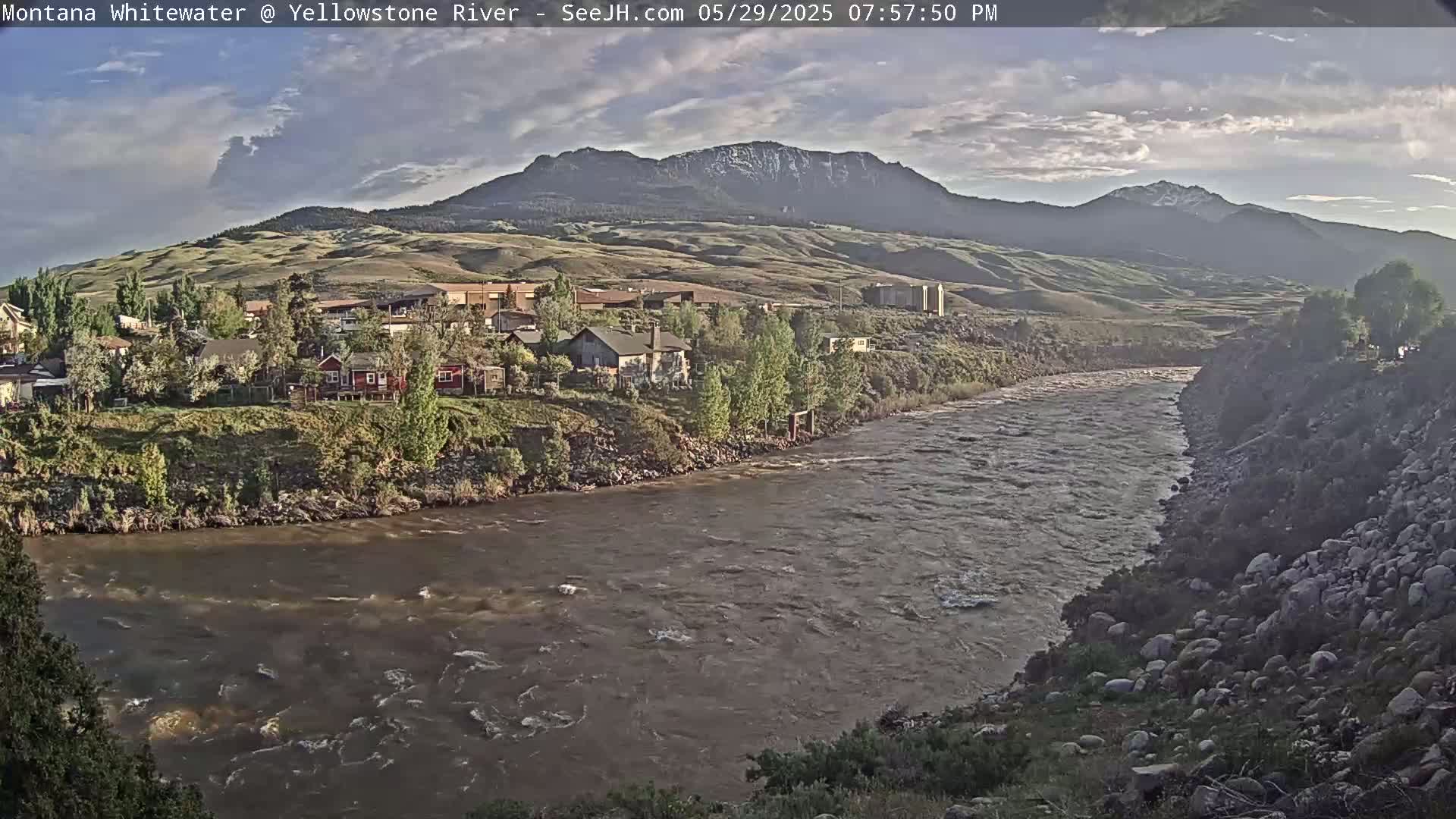 A wide, muddy river flows past a small town nestled at the base of a mountain range under a partly cloudy sky.