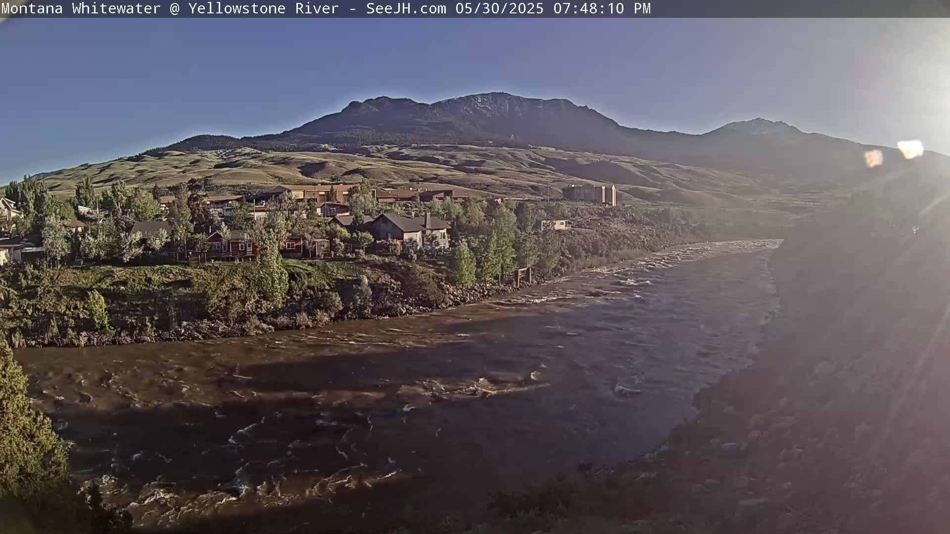 A wide, brown river flows past a small town nestled at the base of a mountain range under a clear, bright sky.
