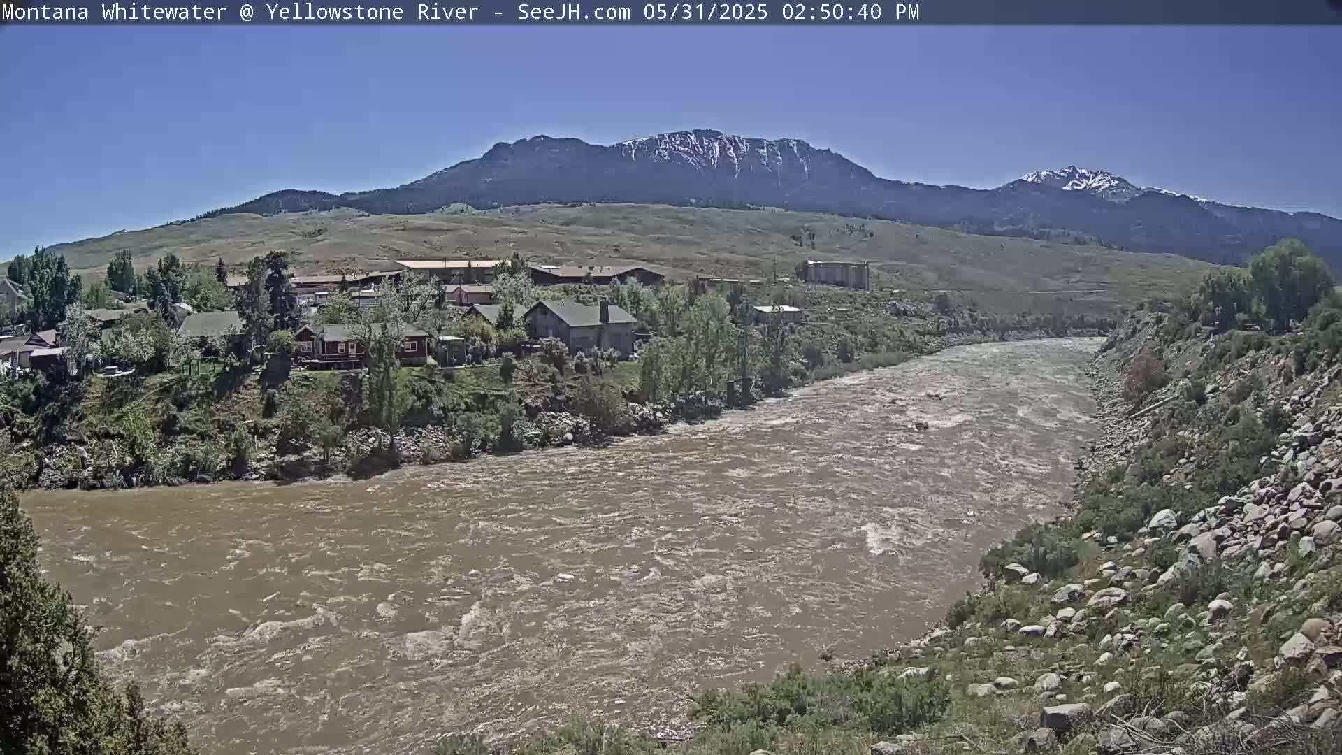 A wide, muddy river flows past a small town nestled at the base of a mountain range under a clear sky.