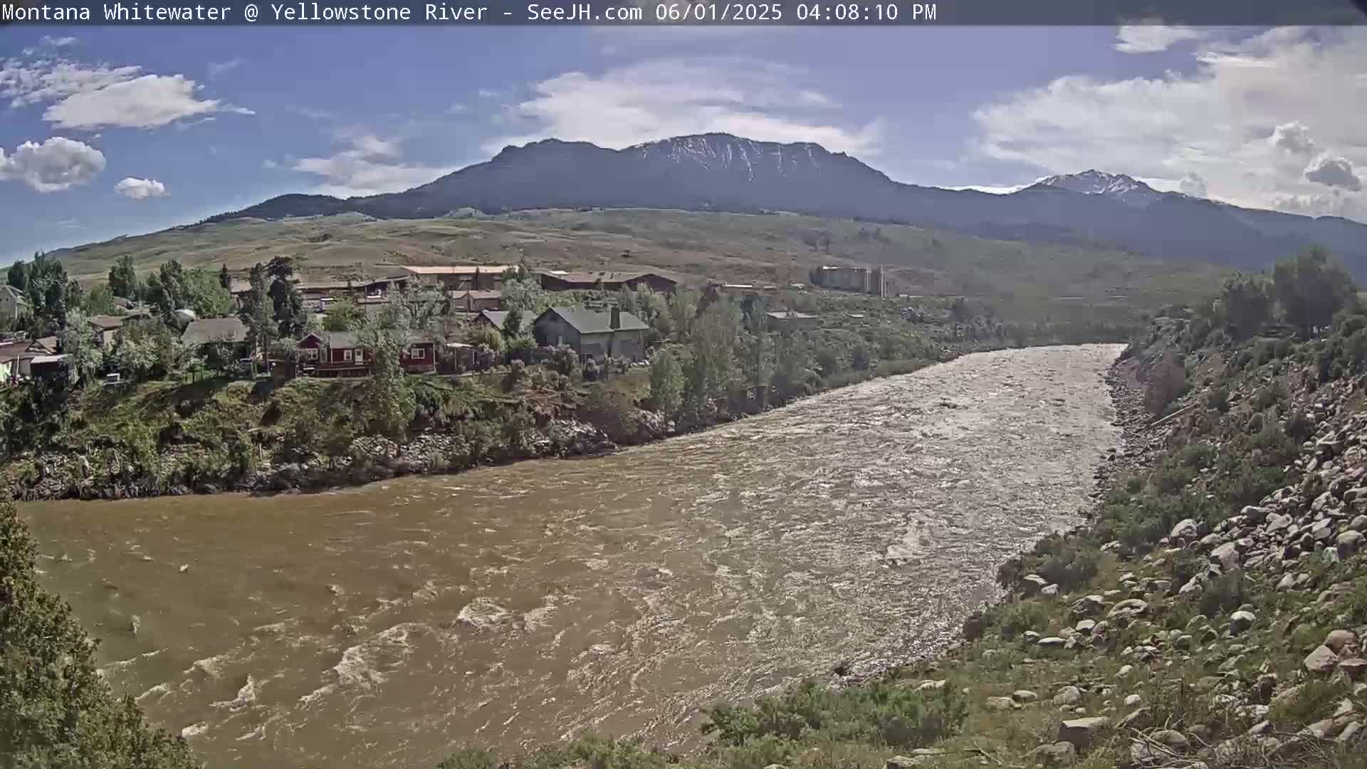 A muddy river flows past a small town nestled beside a mountain range under a partly cloudy sky.