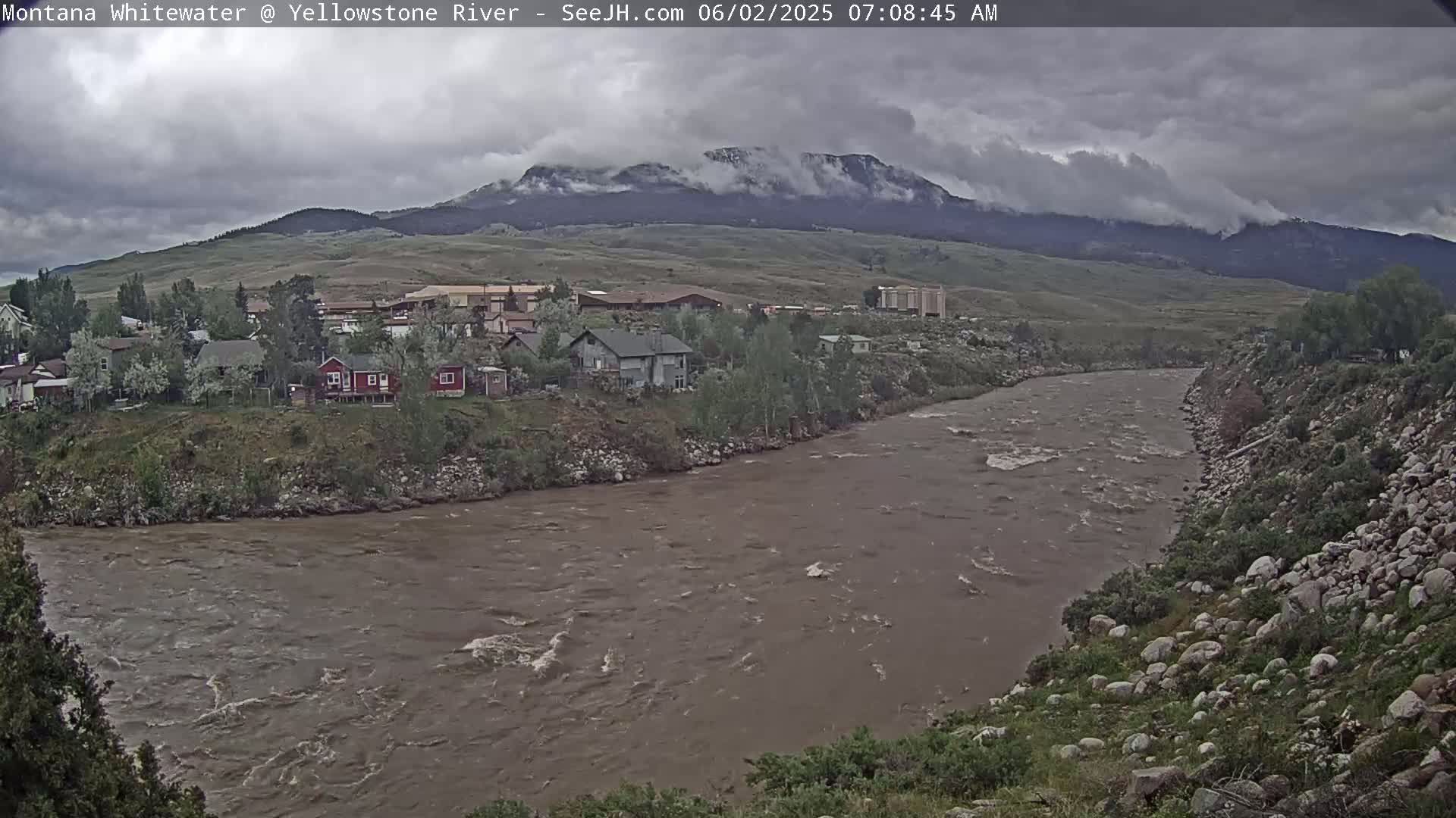 A muddy river flows beneath an overcast sky past a small town nestled at the base of a mountain range.