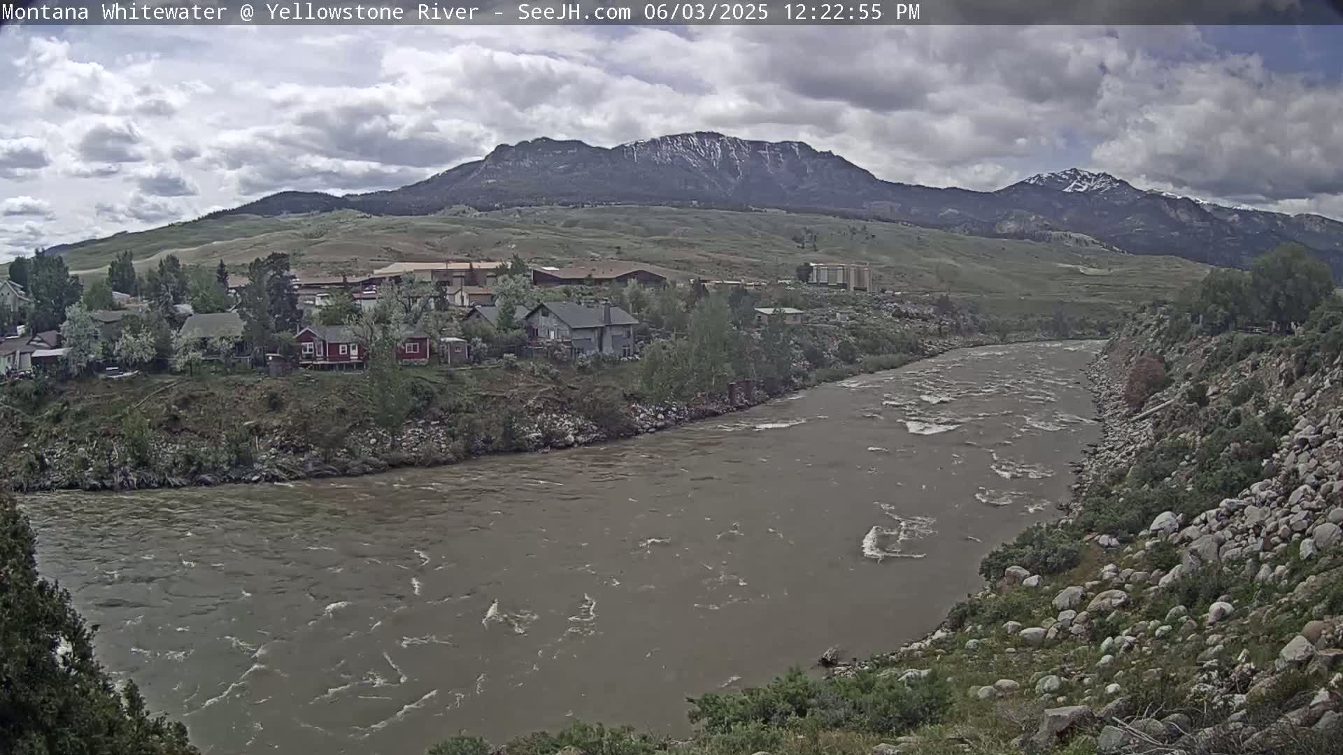 A wide, muddy river flows past a small town nestled at the base of a snow-capped mountain range under a cloudy sky.