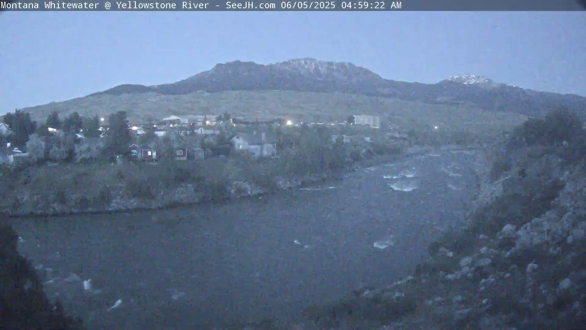 A fast-flowing river winds through a town nestled at the base of a mountain range under a twilight sky.