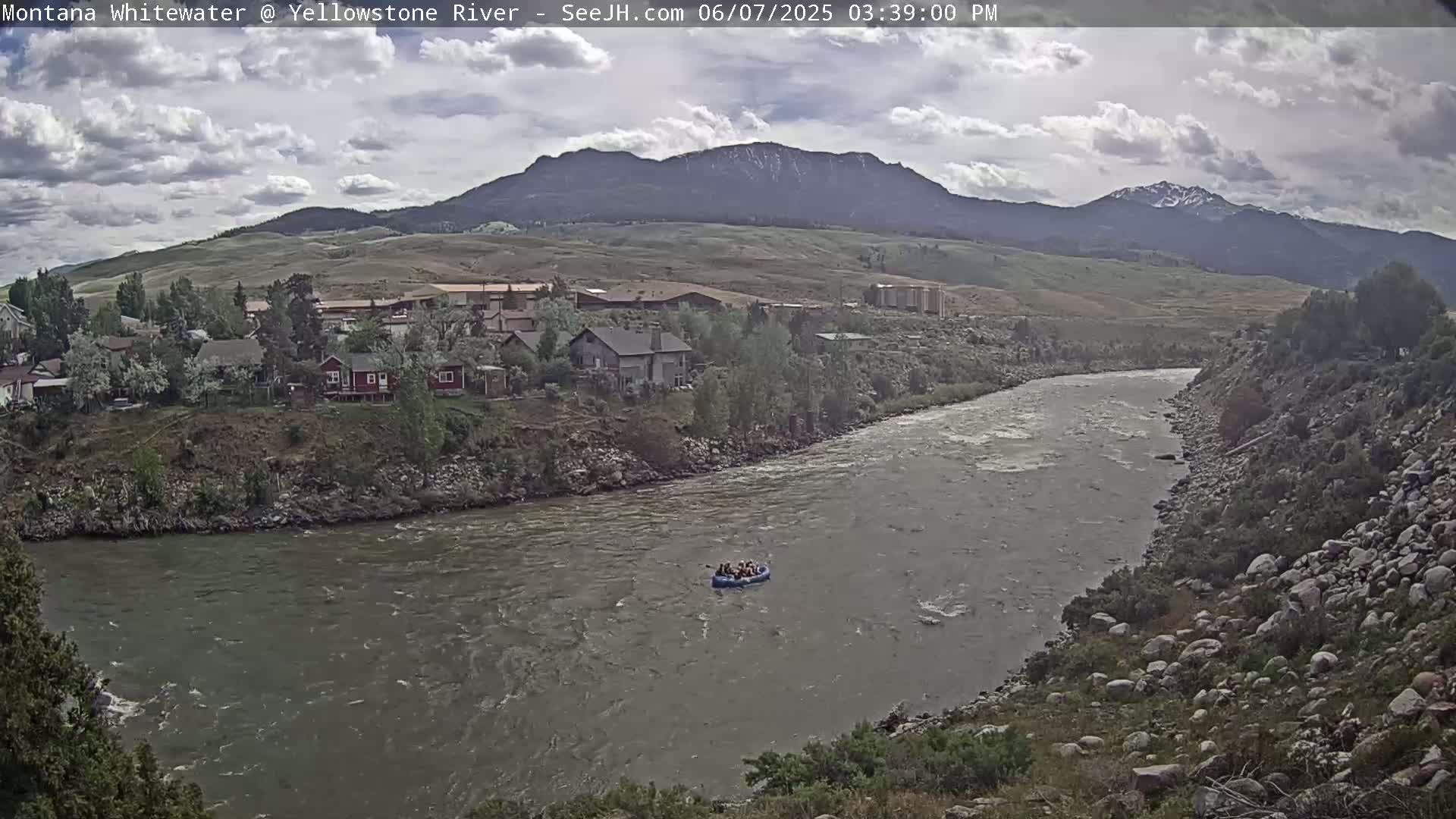 A raft carrying several people floats down a swiftly moving river under a partly cloudy sky, with houses and mountains in the background.