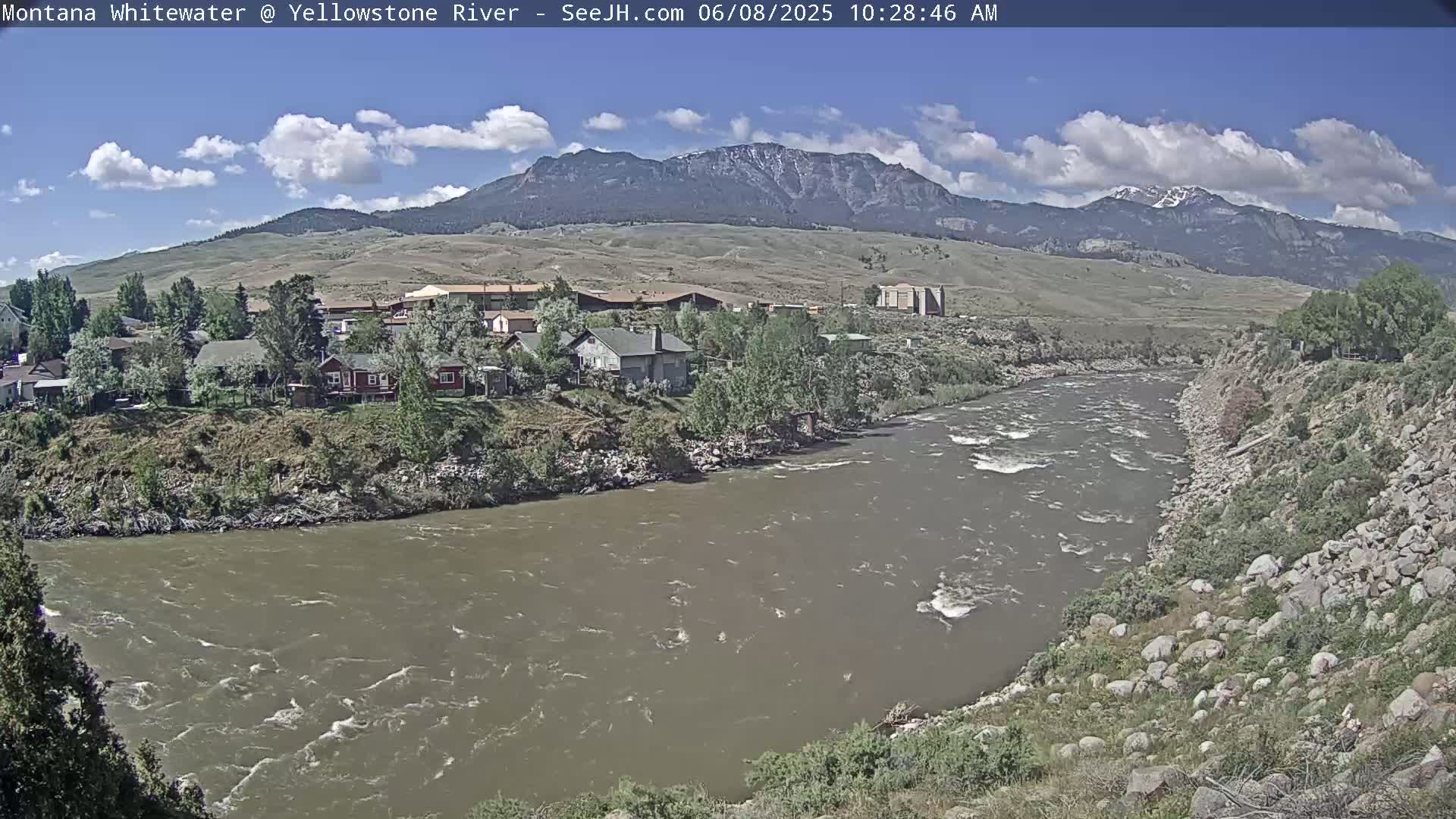 A partly cloudy day reveals a wide, muddy river flowing past a small town nestled against a mountain range with snow-capped peaks.