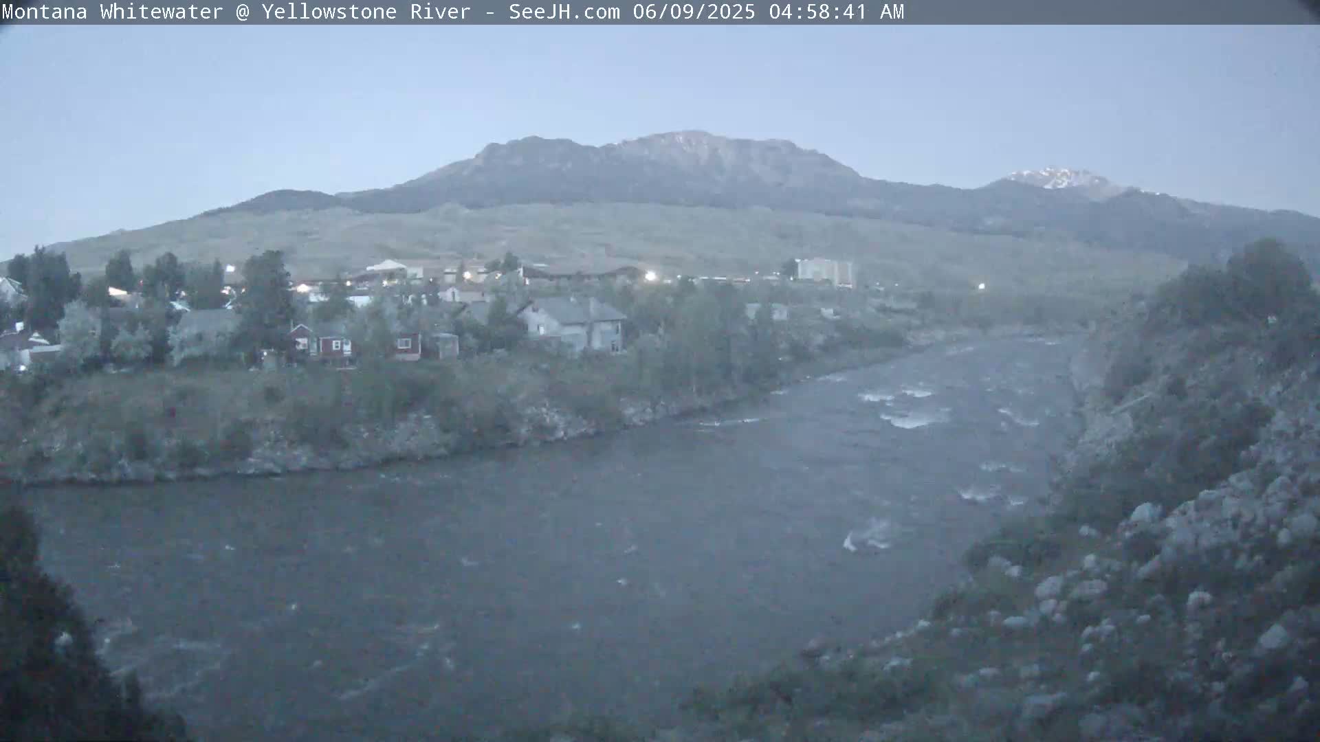 A wide shot shows a river flowing past a town nestled at the base of a mountain range under a twilight sky.