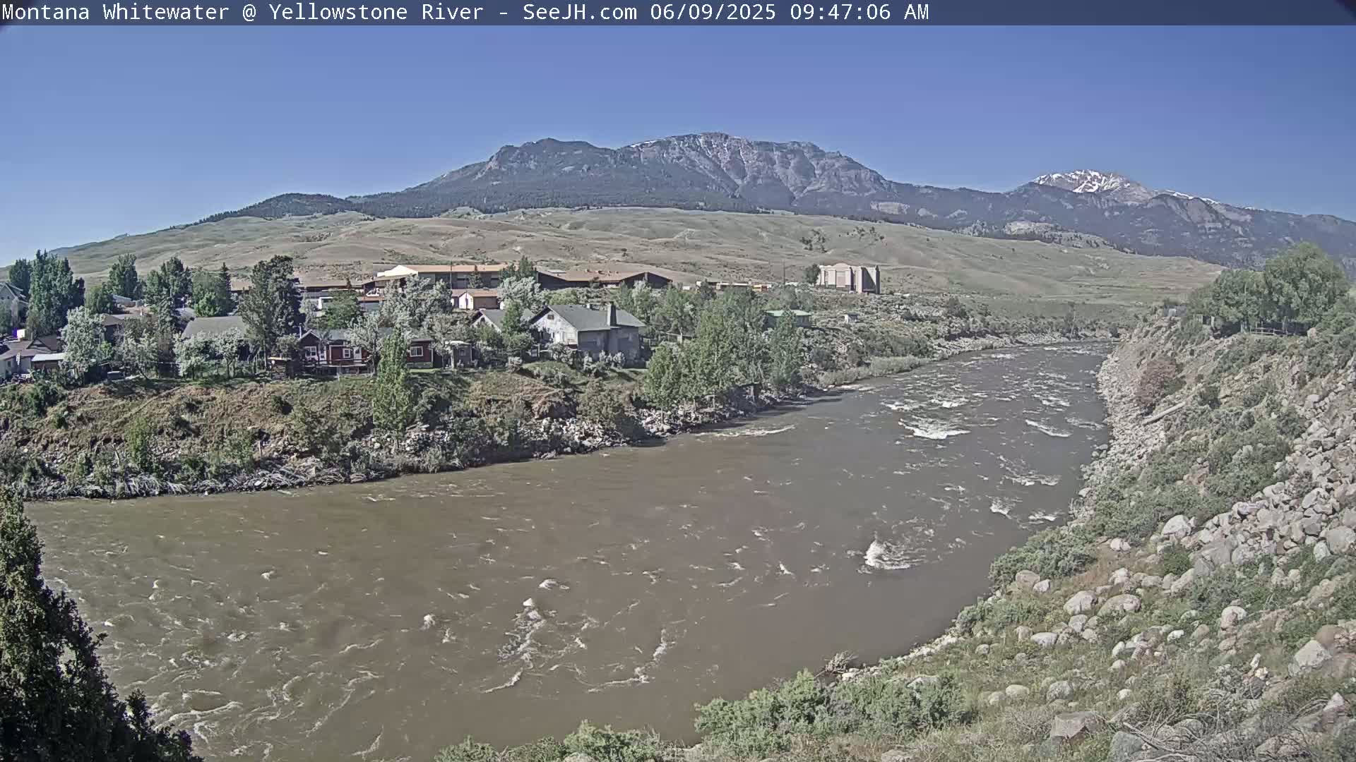 A muddy river flows past a small town nestled against a mountain range under a clear sky.