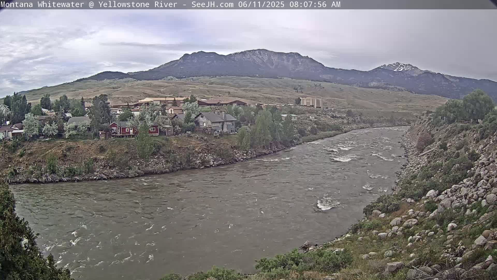 A swiftly flowing river curves past a small town nestled at the base of a mountain range under an overcast sky.