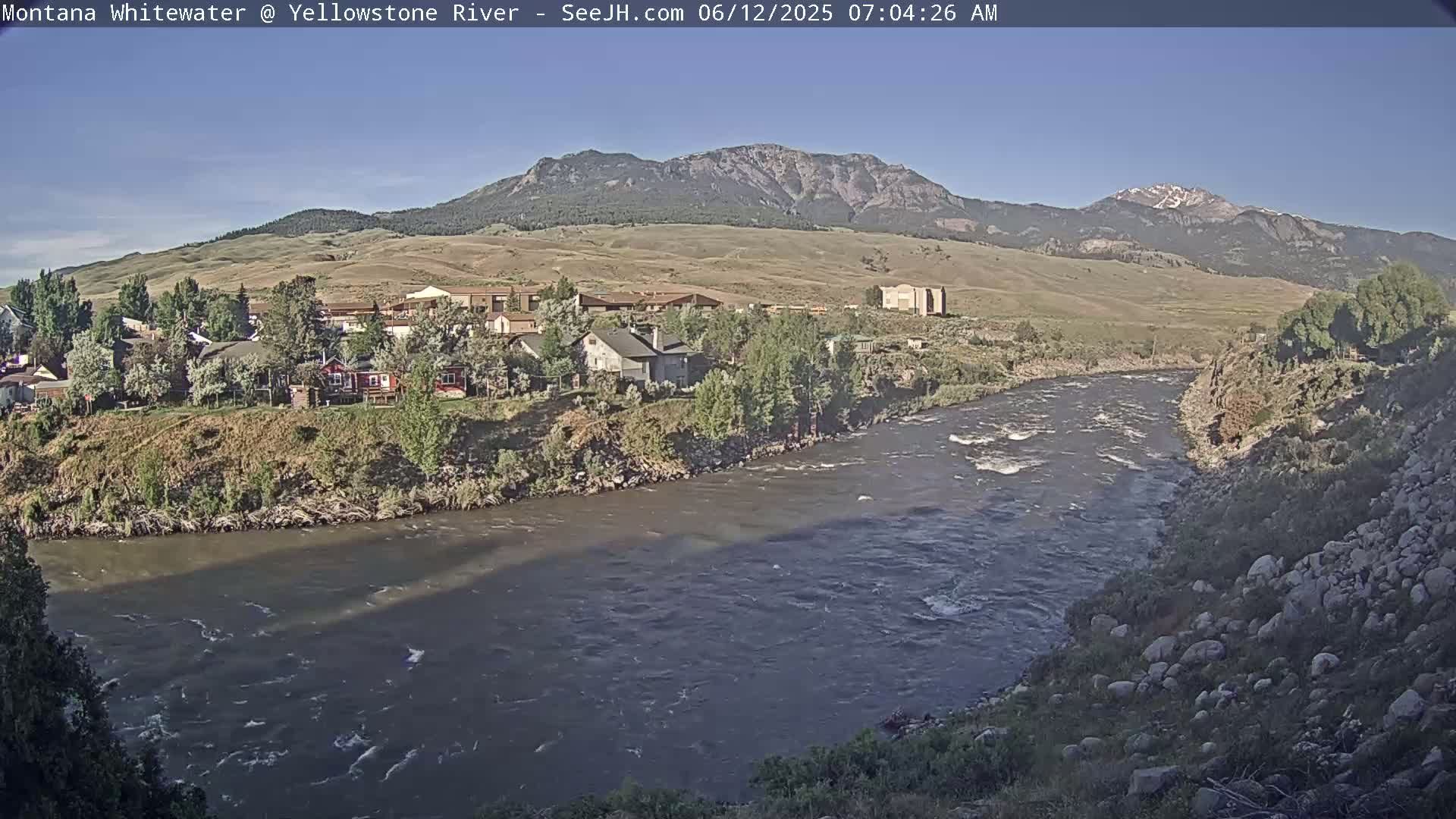 A river flows through a town nestled at the base of a mountain range under a clear sky.