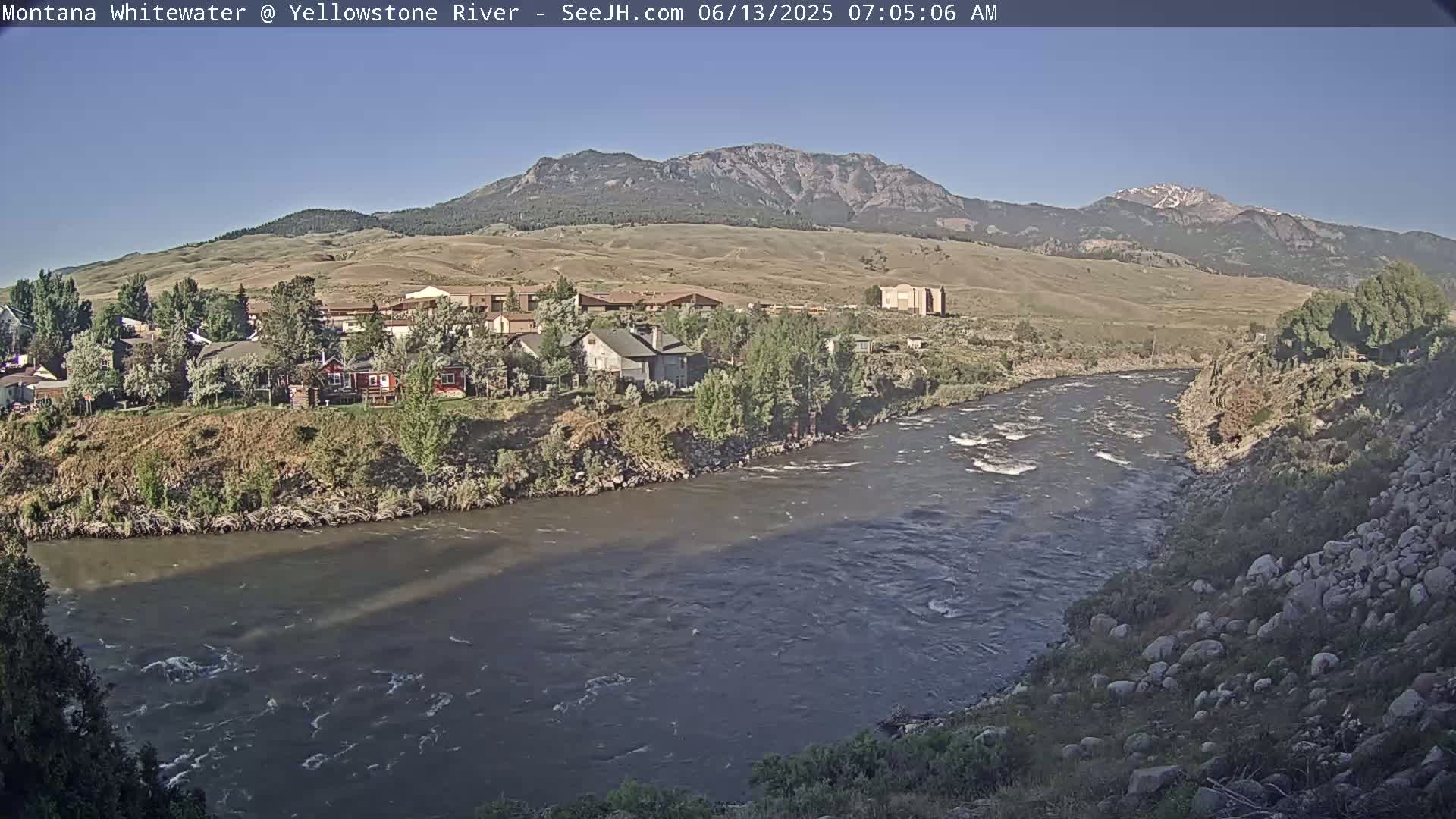 A river flows past a small town nestled at the base of a mountain range under a clear sky.