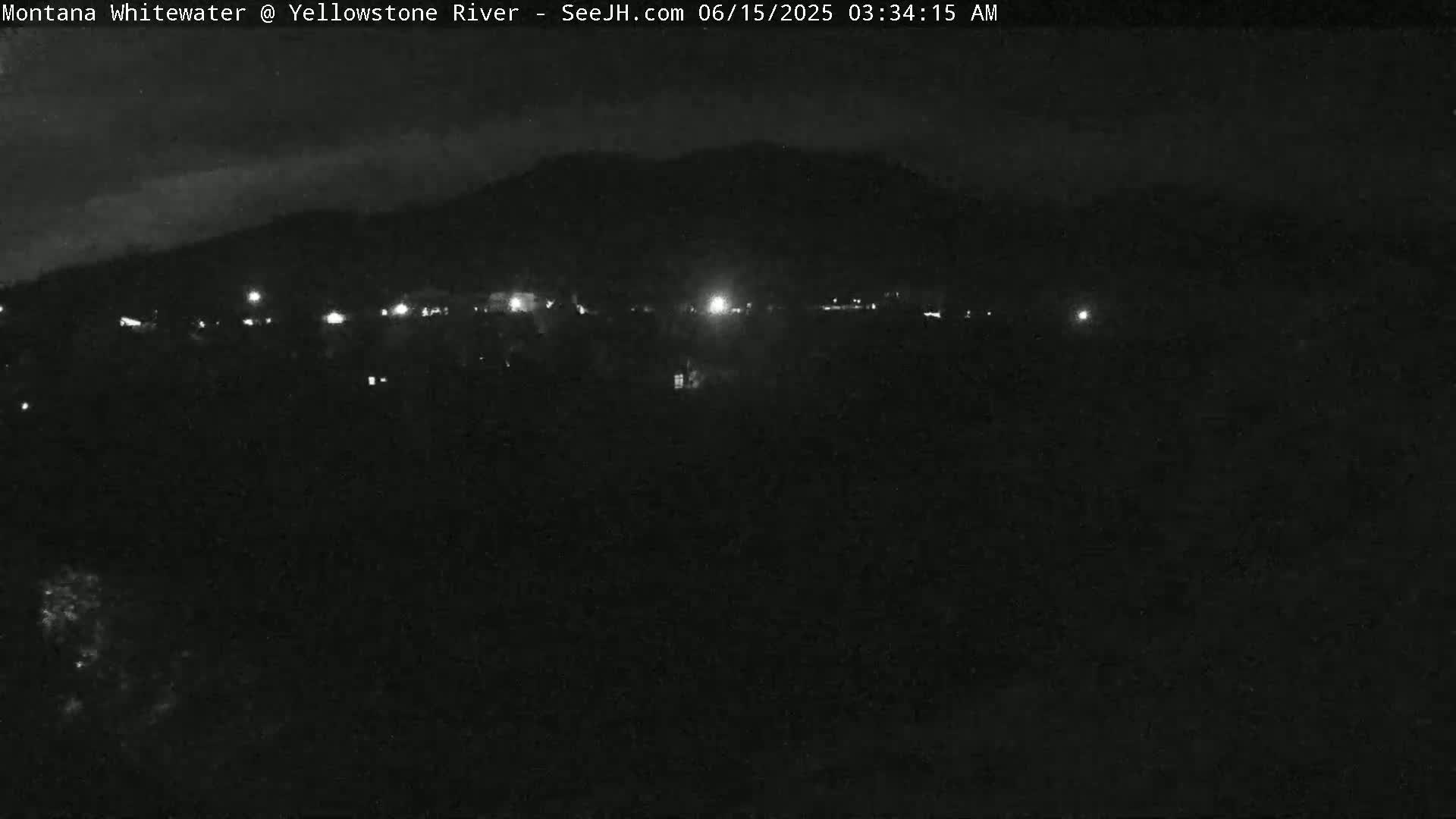A nighttime, dark view shows a distant line of lights at the base of a mountain under a mostly clear sky.