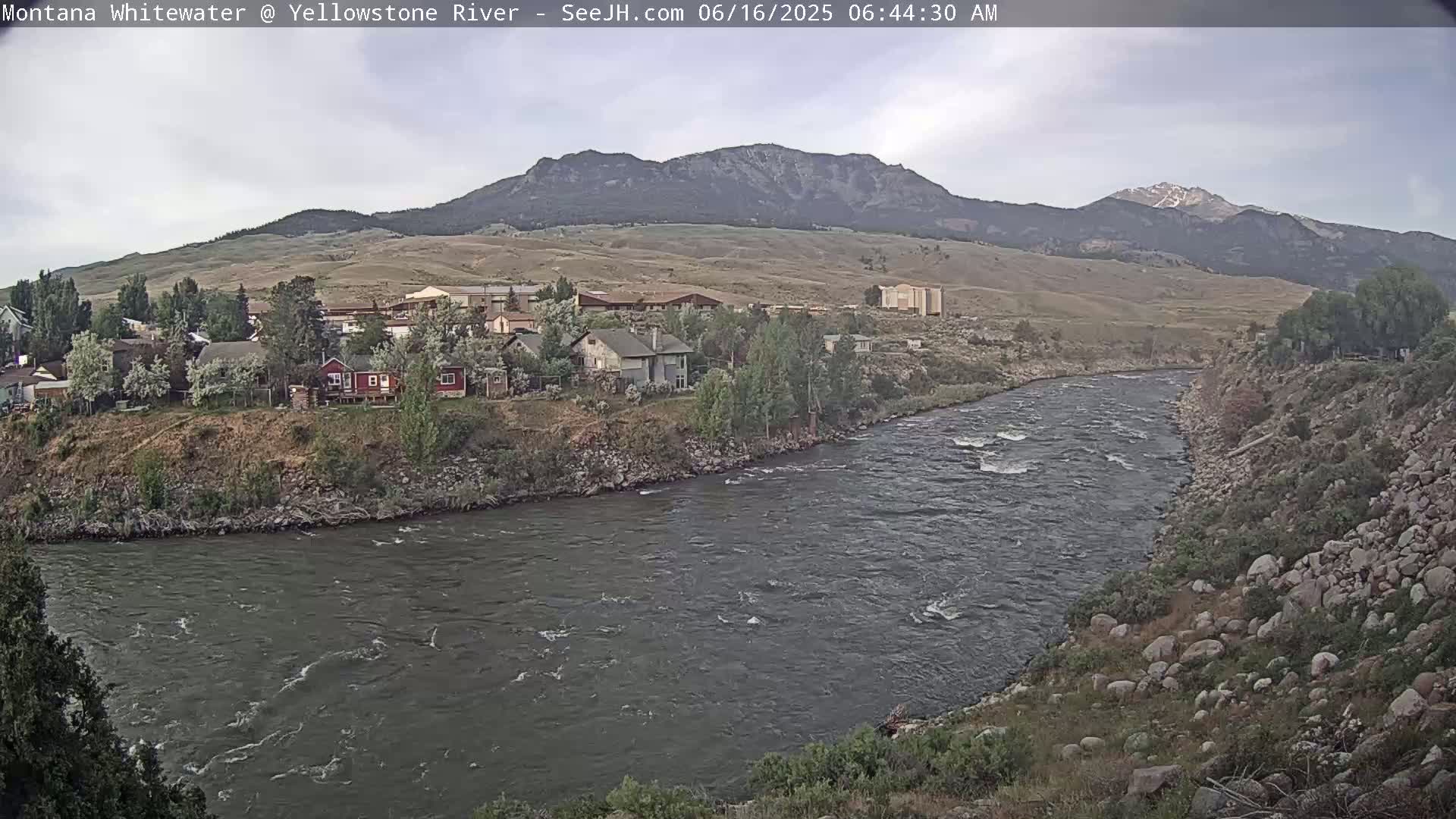 A wide, swiftly flowing river curves past a small town nestled against a hillside beneath a partly cloudy sky and mountains in the distance.