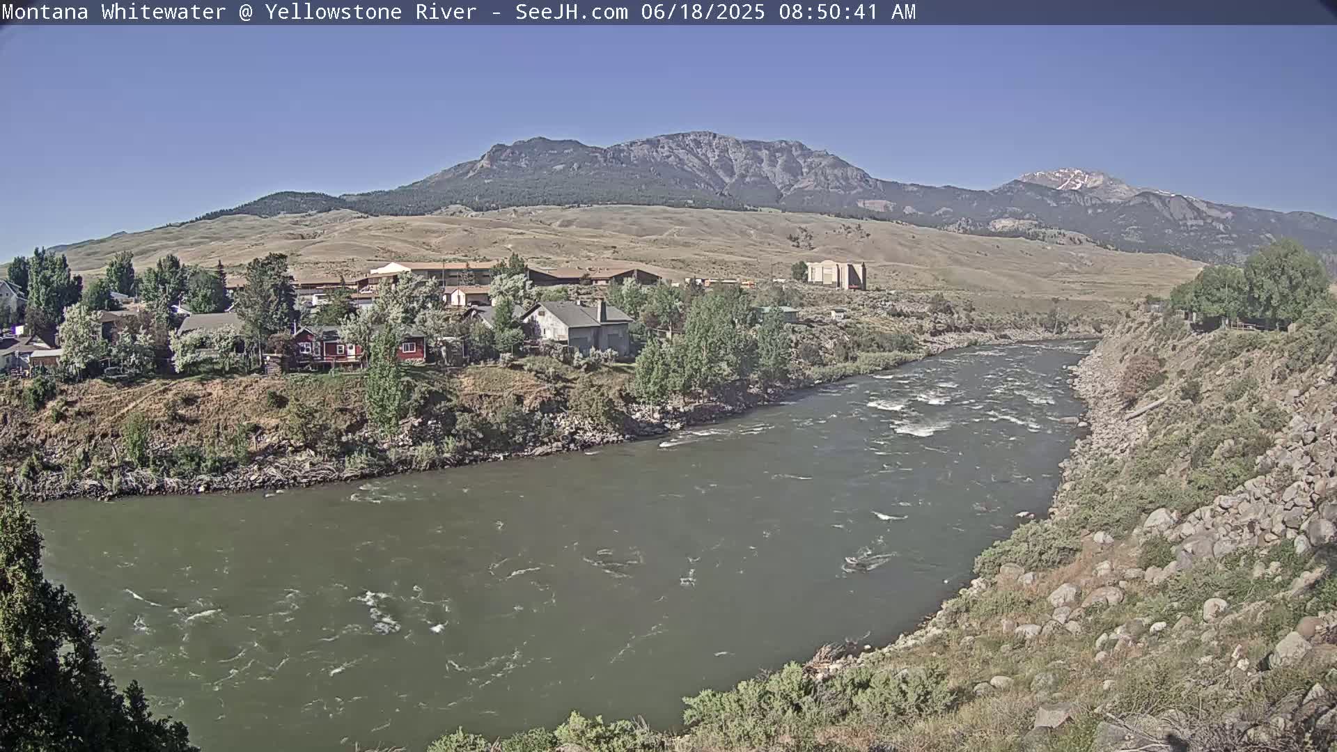 A swiftly flowing river bends around a small town nestled against a mountain range under a clear, sunny sky.