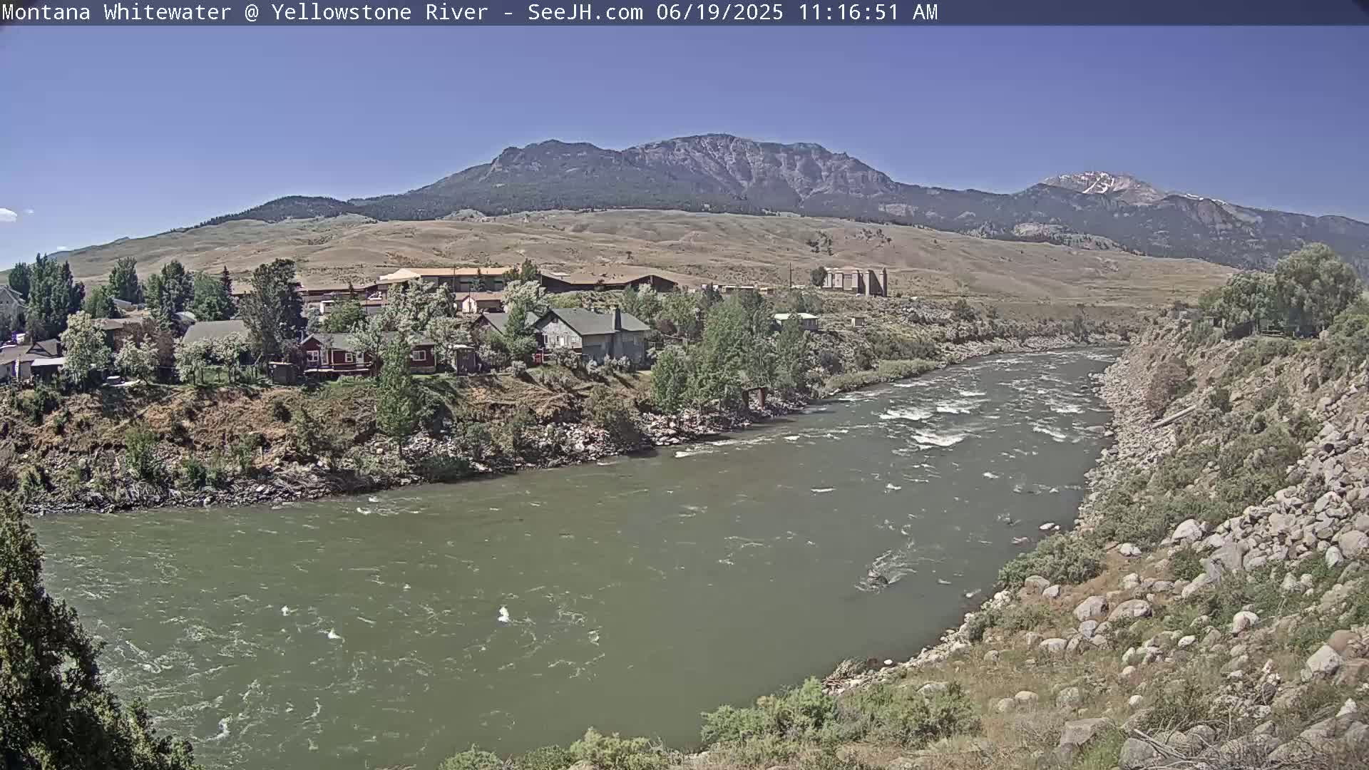 A swiftly flowing river winds through a valley under a clear blue sky, past a small town nestled against a mountain range.
