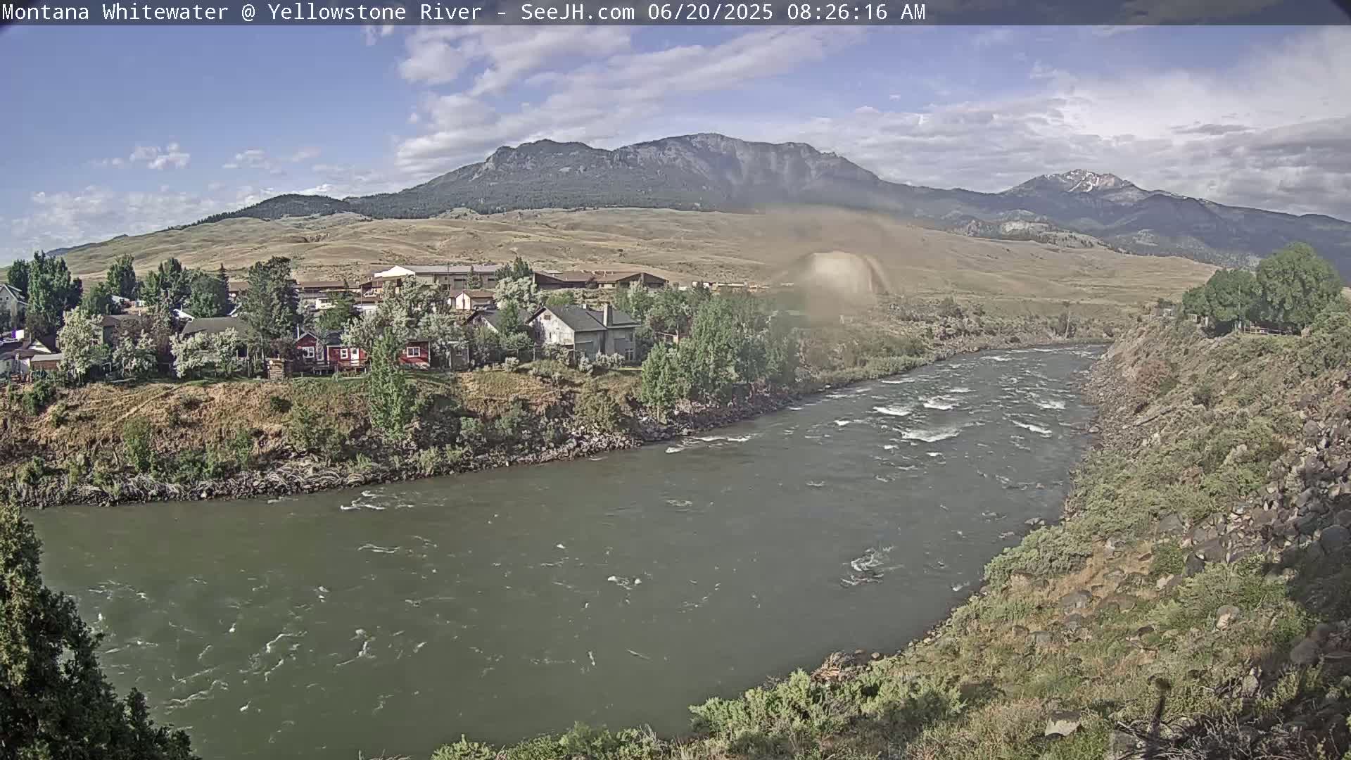 A partly cloudy day reveals a flowing river snaking through a valley of green hills and houses, with mountains in the background.
