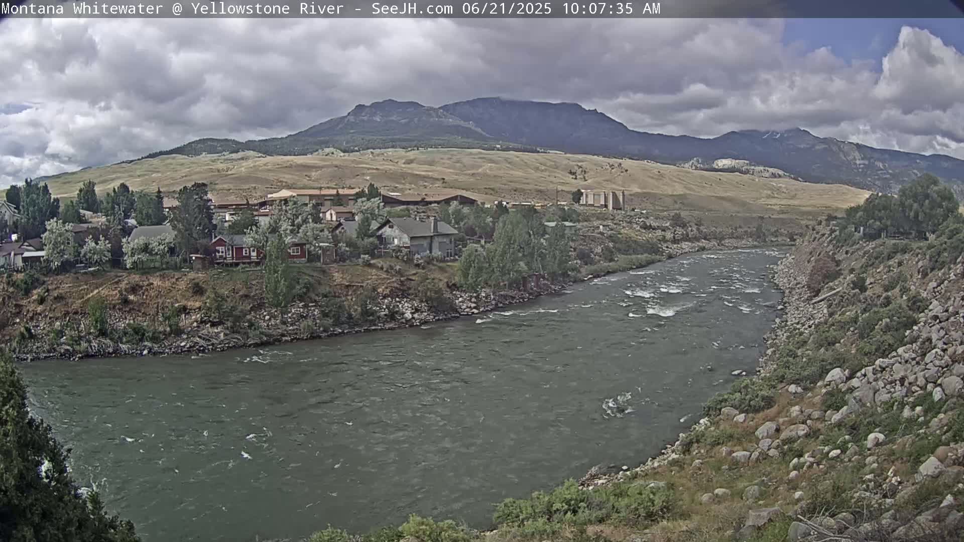 A partly cloudy day shows a rocky river flowing past a small town nestled against a mountain range.