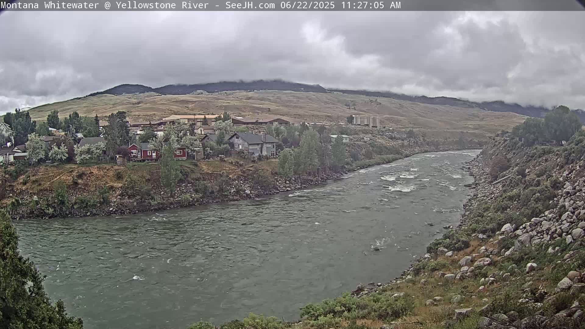 A wide, rocky river flows past a small town nestled against a hillside under a cloudy sky.