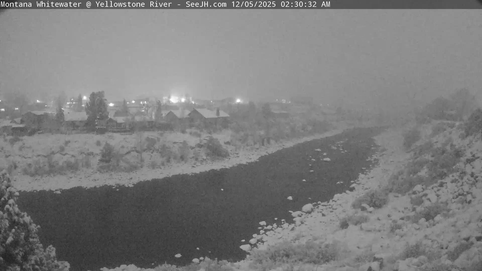 A snowy winter night scene reveals a dark river flowing past snow-covered banks and distant houses with glowing lights, all under conditions of active snowfall and low visibility.