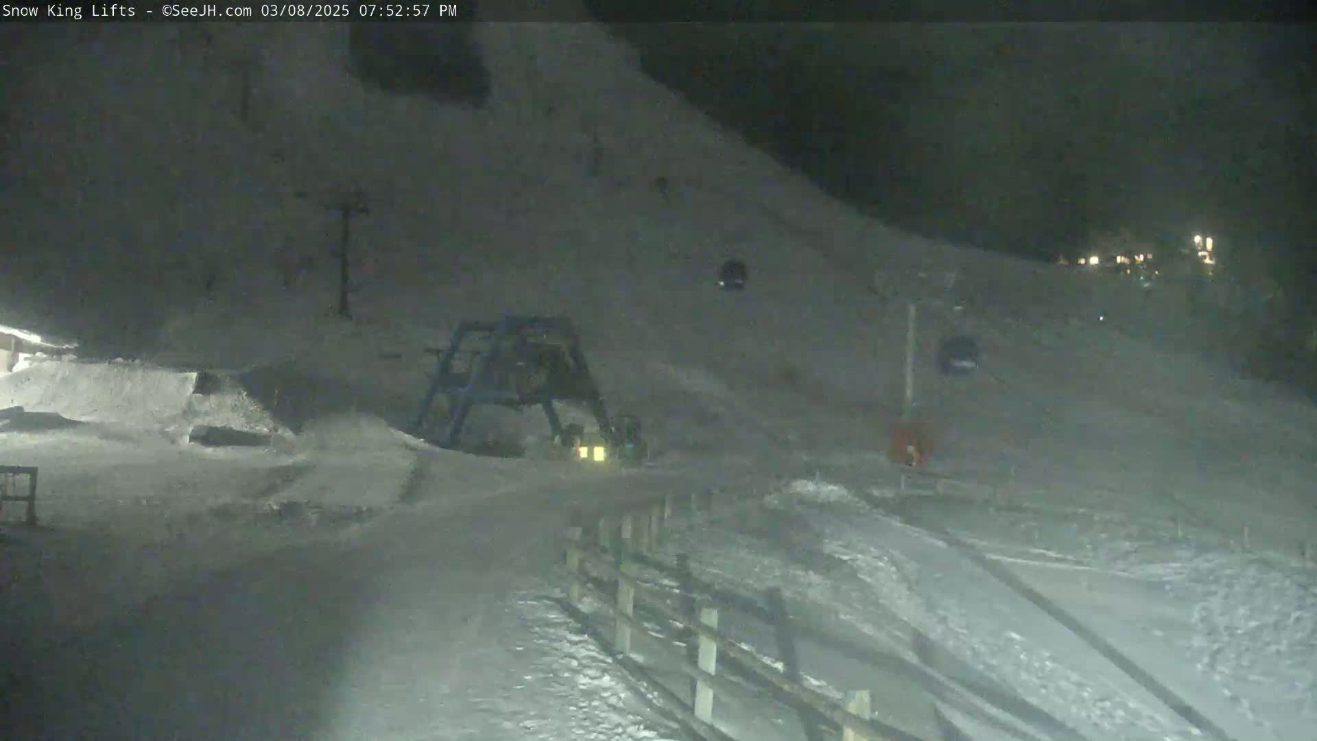A nighttime view of a snow-covered ski slope with a ski lift and snow-grooming equipment, under dark conditions.