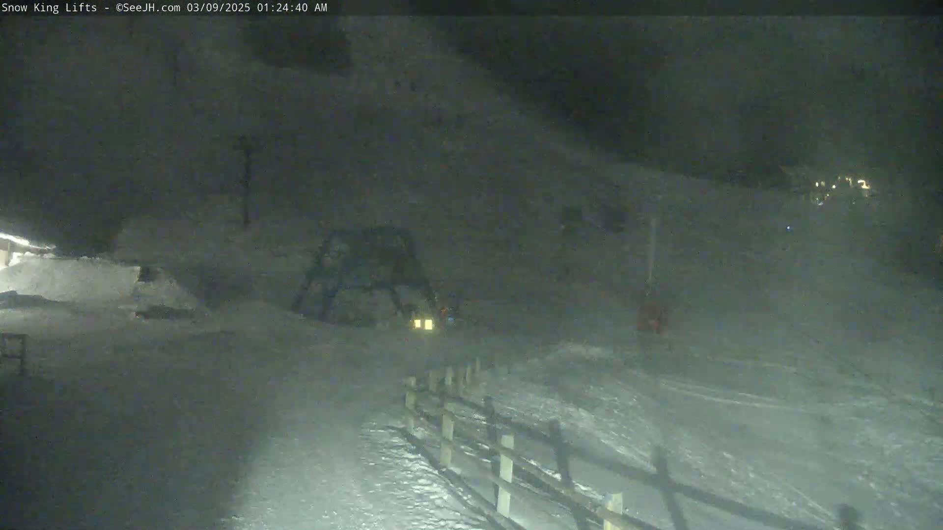 A snow-covered area at night, with a ski lift and a fence visible, under dark, snowy conditions.