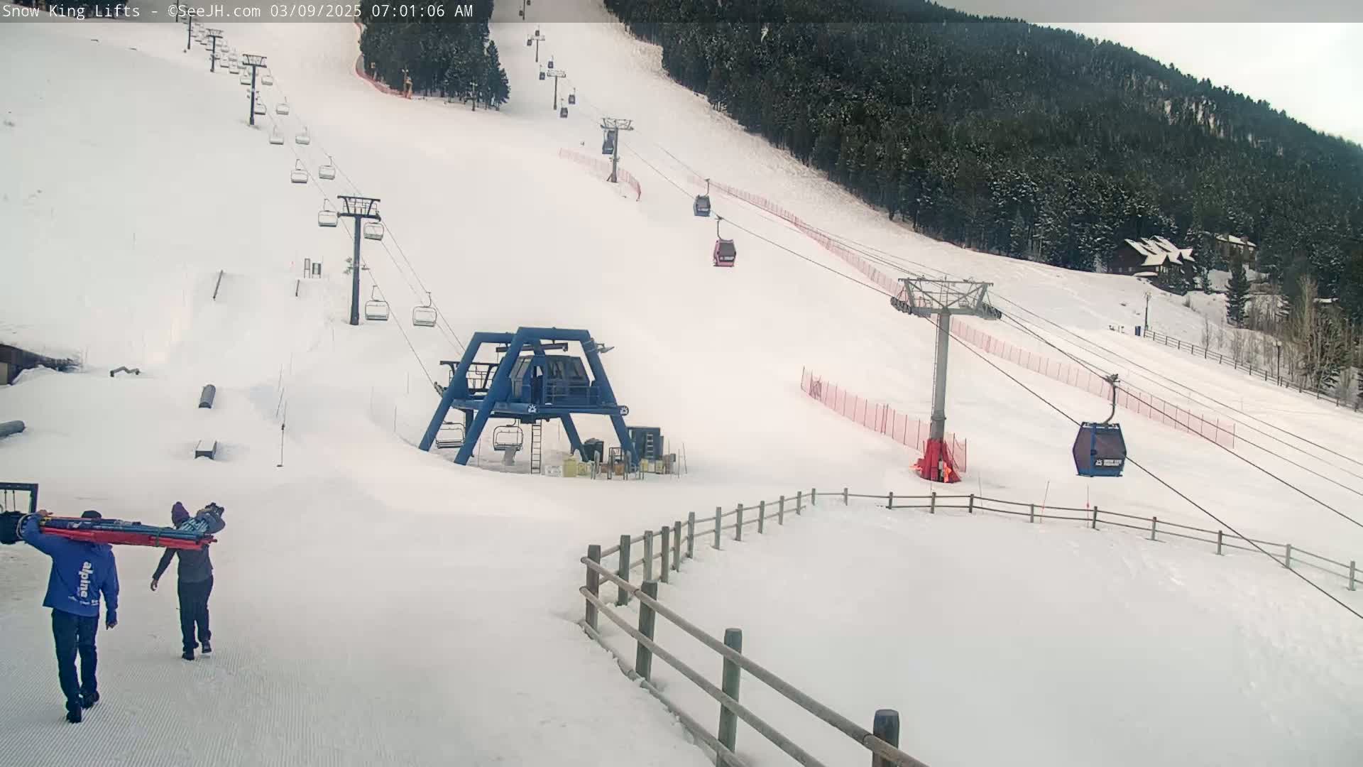 Two people carry a stretcher across a snow-covered ski slope with several chairlifts and gondolas visible, under a cloudy sky.