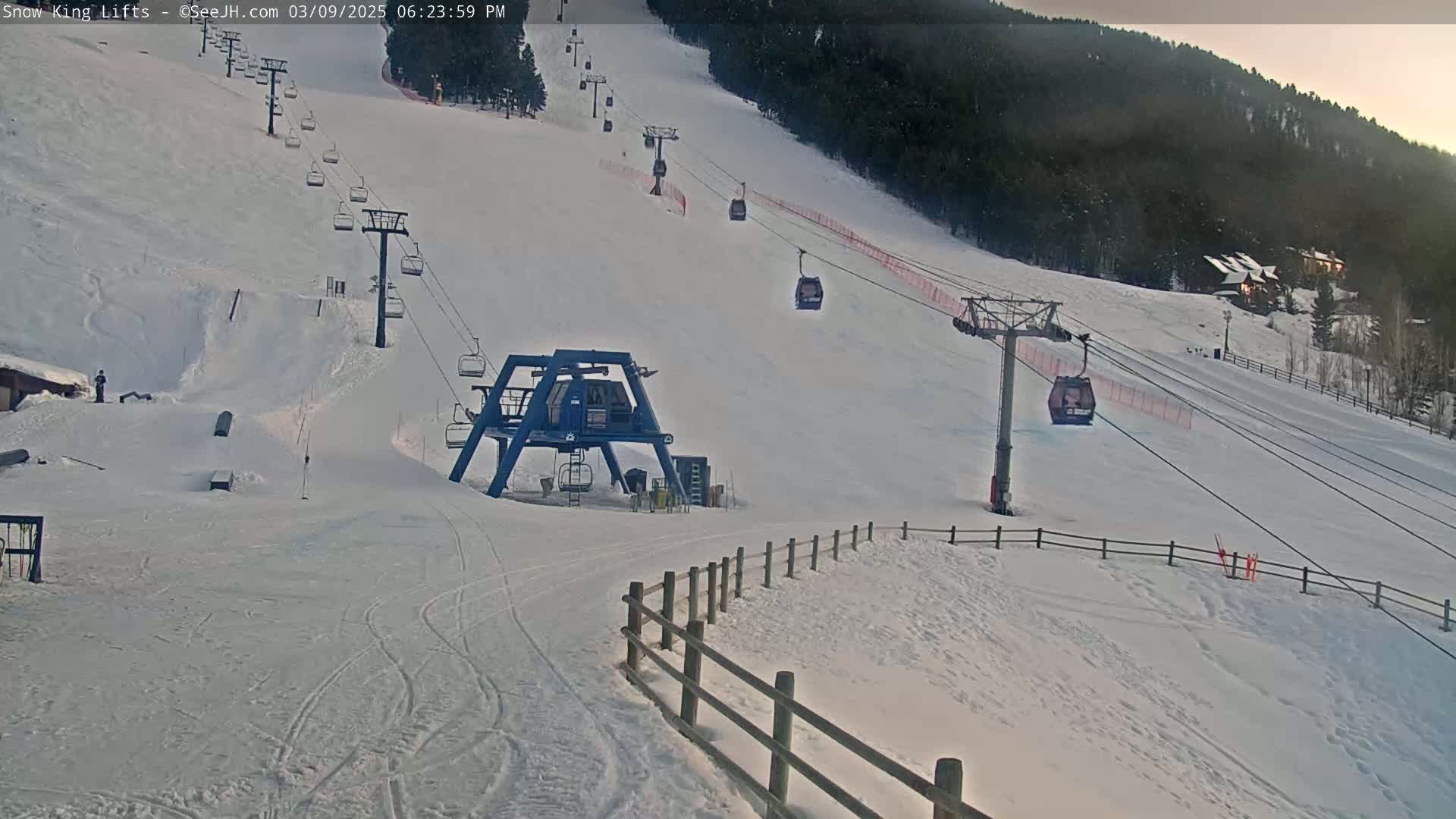 A ski slope covered in snow under a twilight sky shows several chairlifts and gondolas, with a wooden fence running along the bottom.