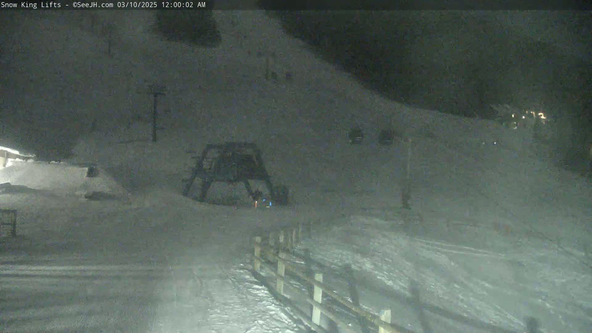 A nighttime view of a snow-covered ski slope with a ski lift and a fence, under dark conditions.