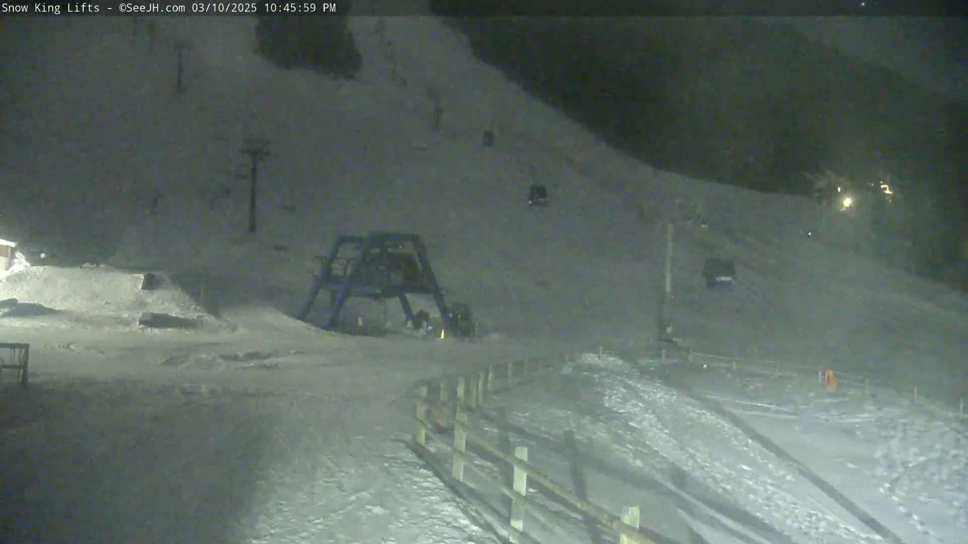 A snow-covered ski slope at night is illuminated, showing a ski lift and several vehicles near the base of the hill.