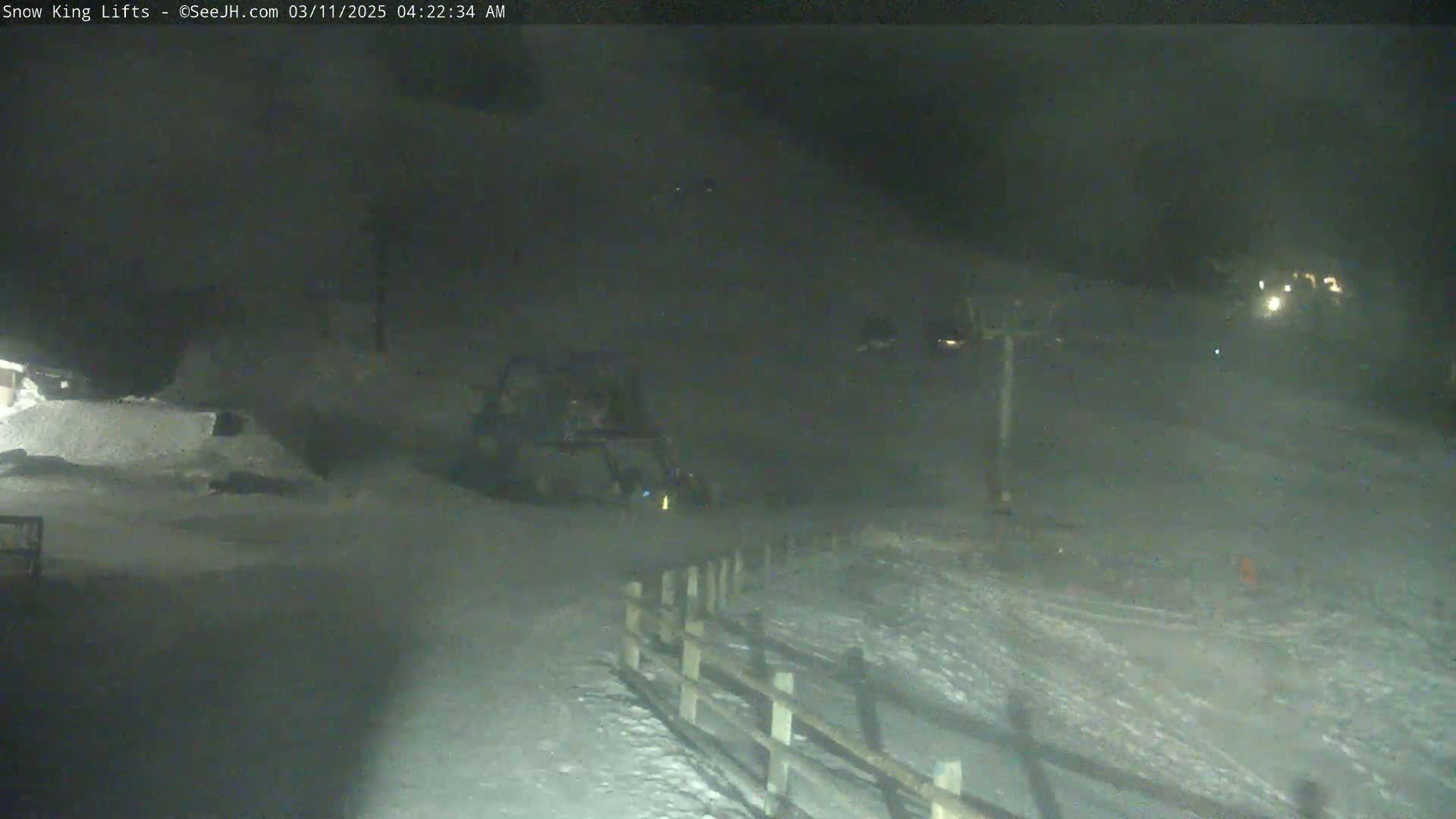 A nighttime, snowy scene shows a snowcat near a wooden fence and a ski lift in the distance under hazy conditions.