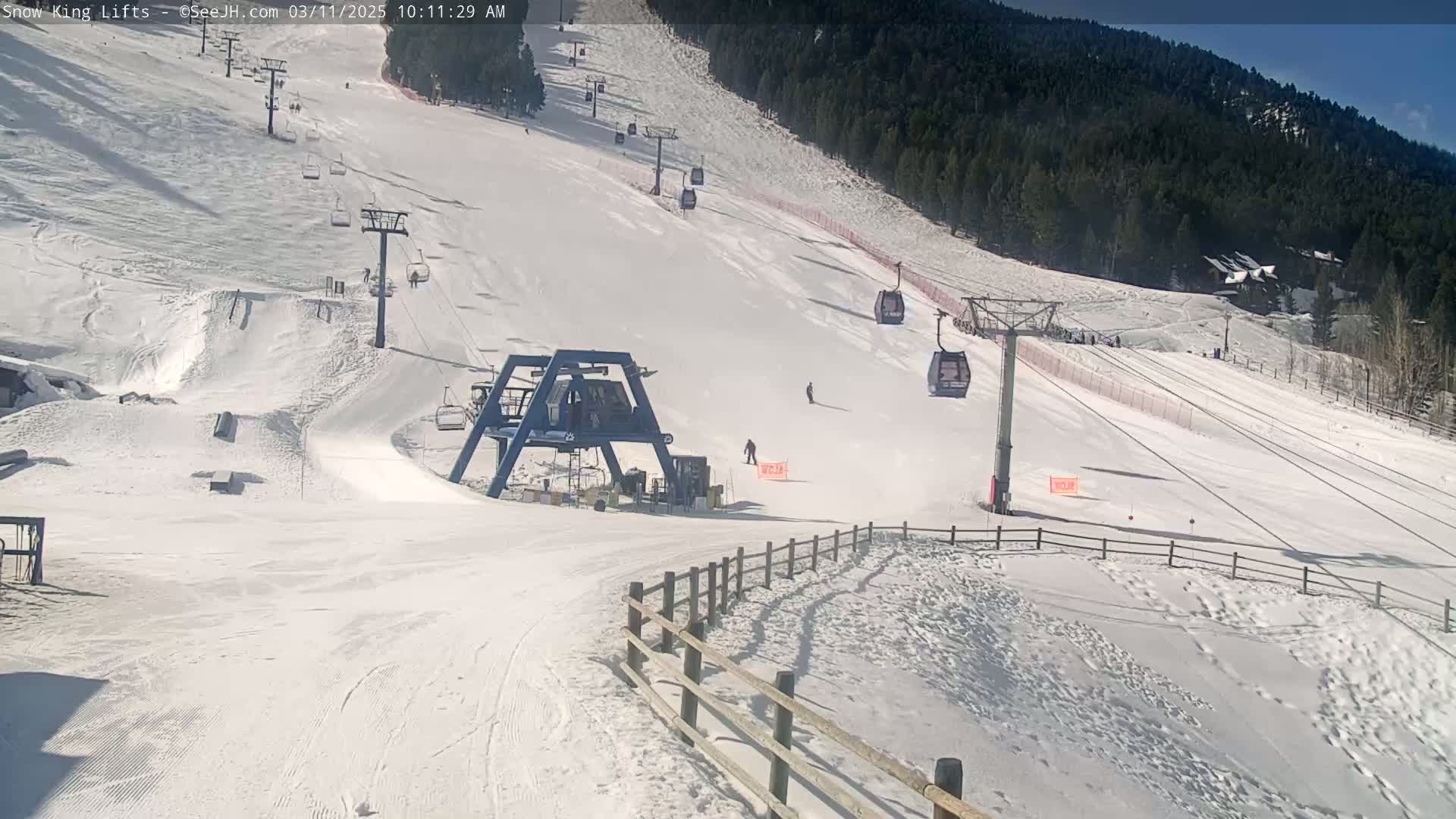 A sunny day at a snow-covered ski resort shows a ski lift and several skiers on a slope with a forested mountain in the background.