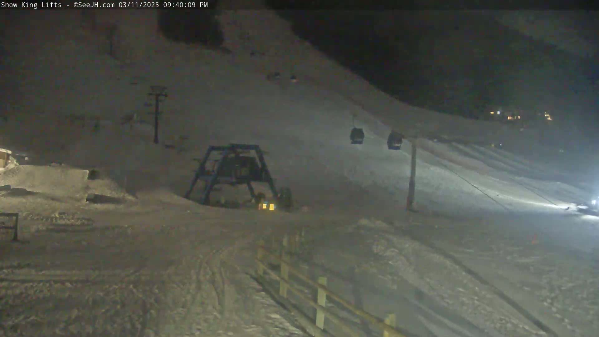 A nighttime view of a snow-covered ski slope with a chairlift and snowmaking equipment visible under dark conditions.