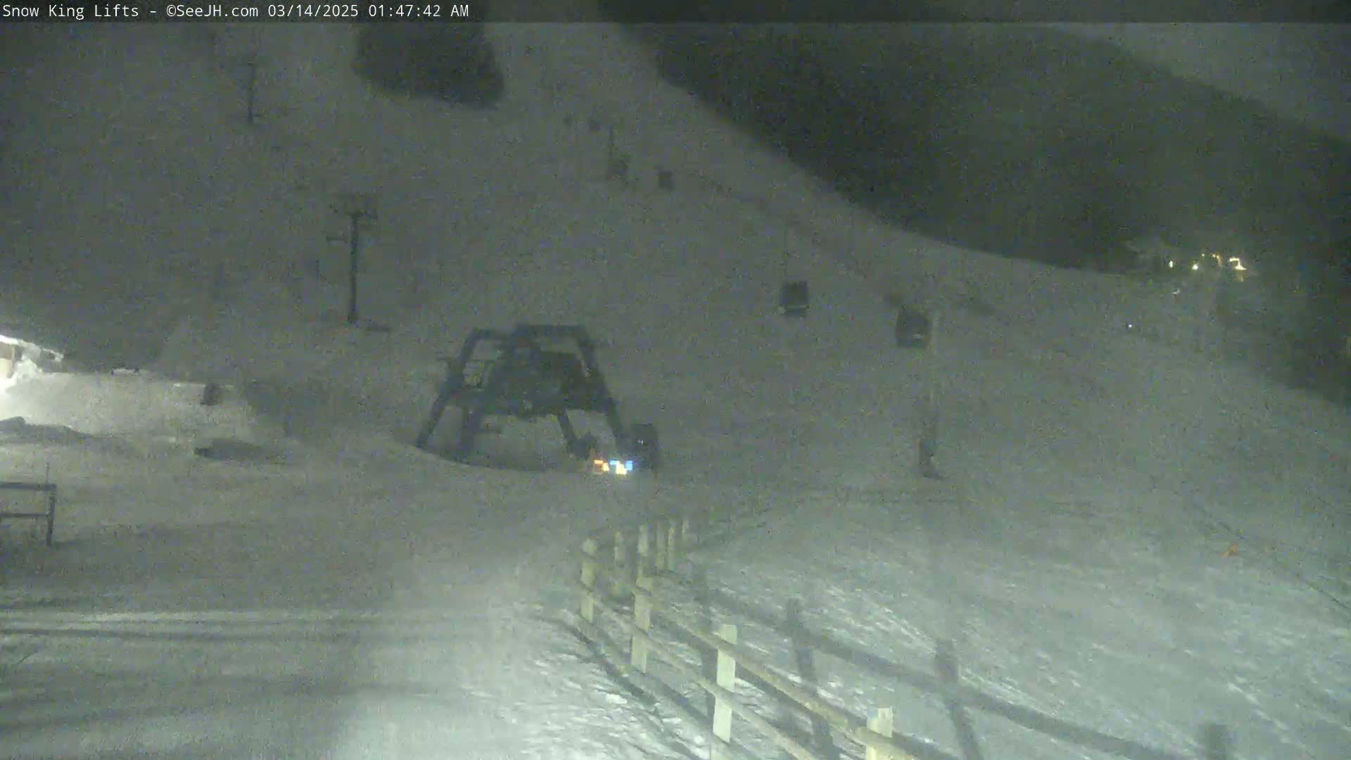 A nighttime, snow-covered ski slope is visible, showing a ski lift and a wooden fence under dark, possibly cloudy conditions.