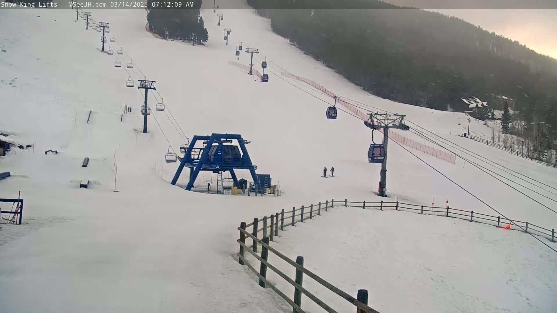 A snow-covered ski slope with several chairlifts and two skiers visible on a somewhat overcast day.