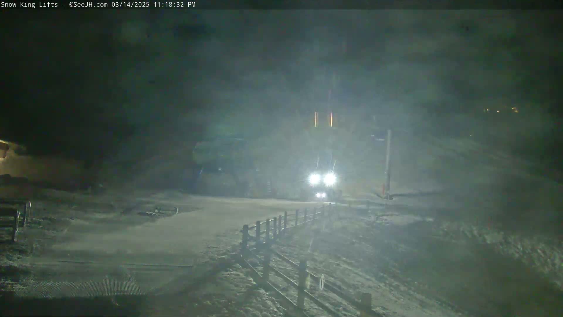 A snow-covered road at night, with a vehicle's headlights visible in the distance, under foggy or snowy conditions.