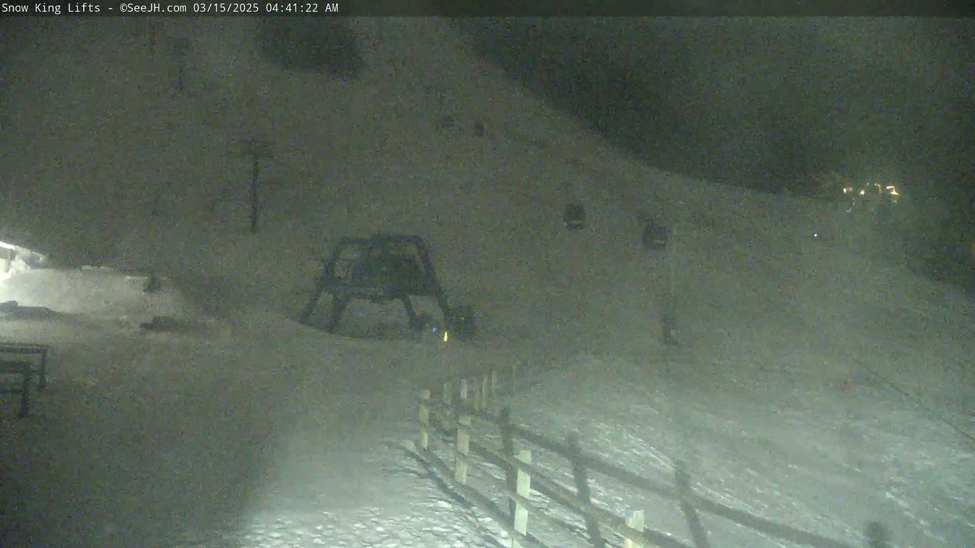 A nighttime, snowy scene shows a ski lift and a snow-covered slope with a wooden fence.