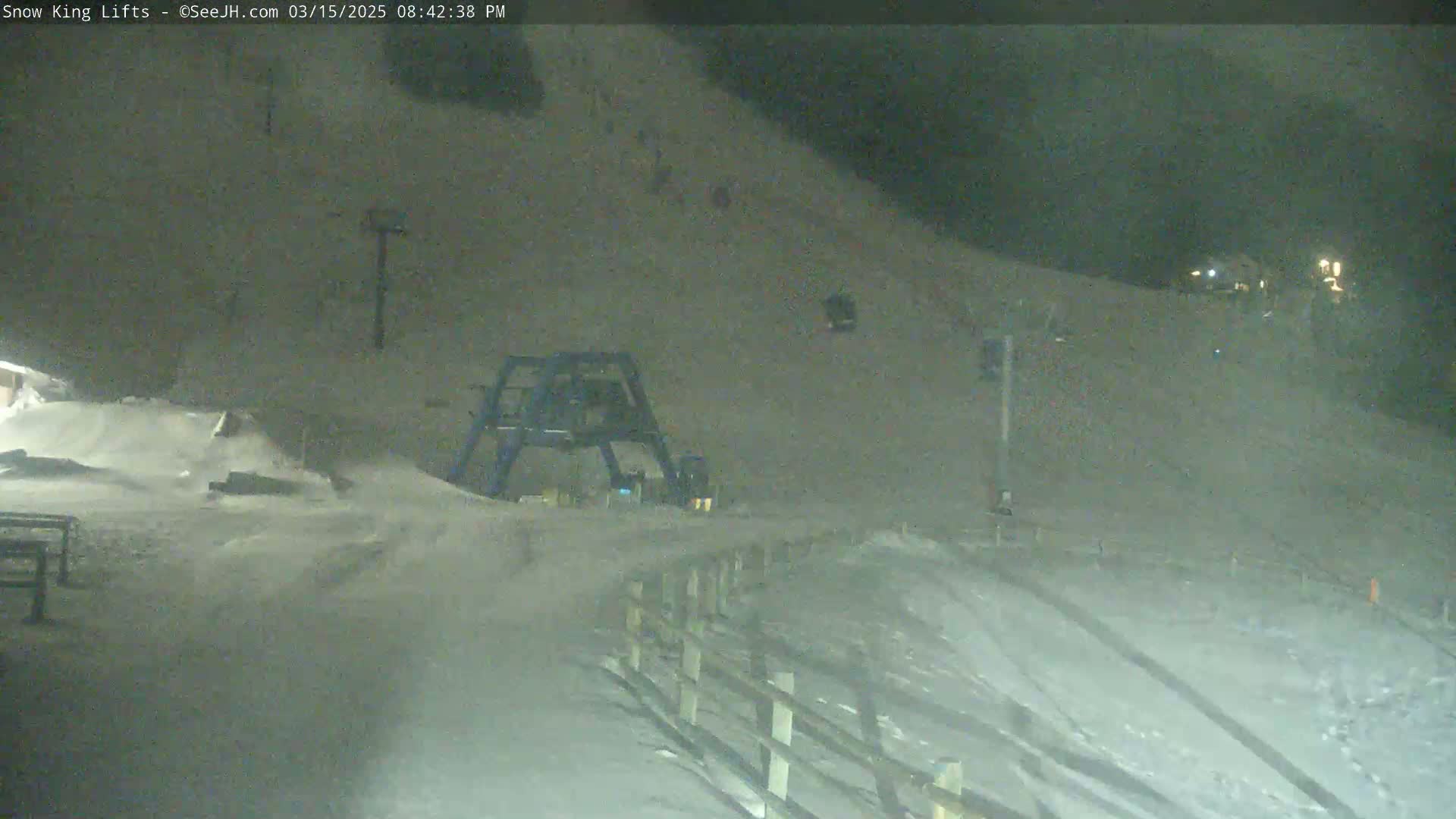 A snow-covered ski slope at night, illuminated by dim lights, features a ski lift and a snow-covered fence.
