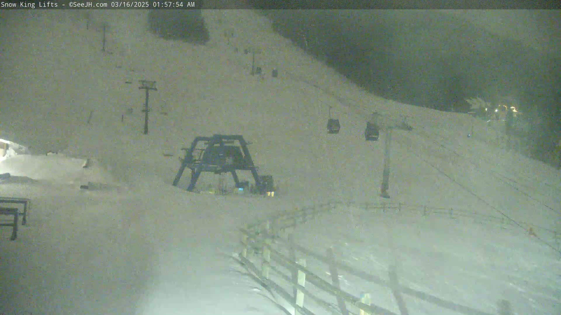 A nighttime view of a snow-covered ski slope with a chairlift and its terminal station, under cloudy conditions.