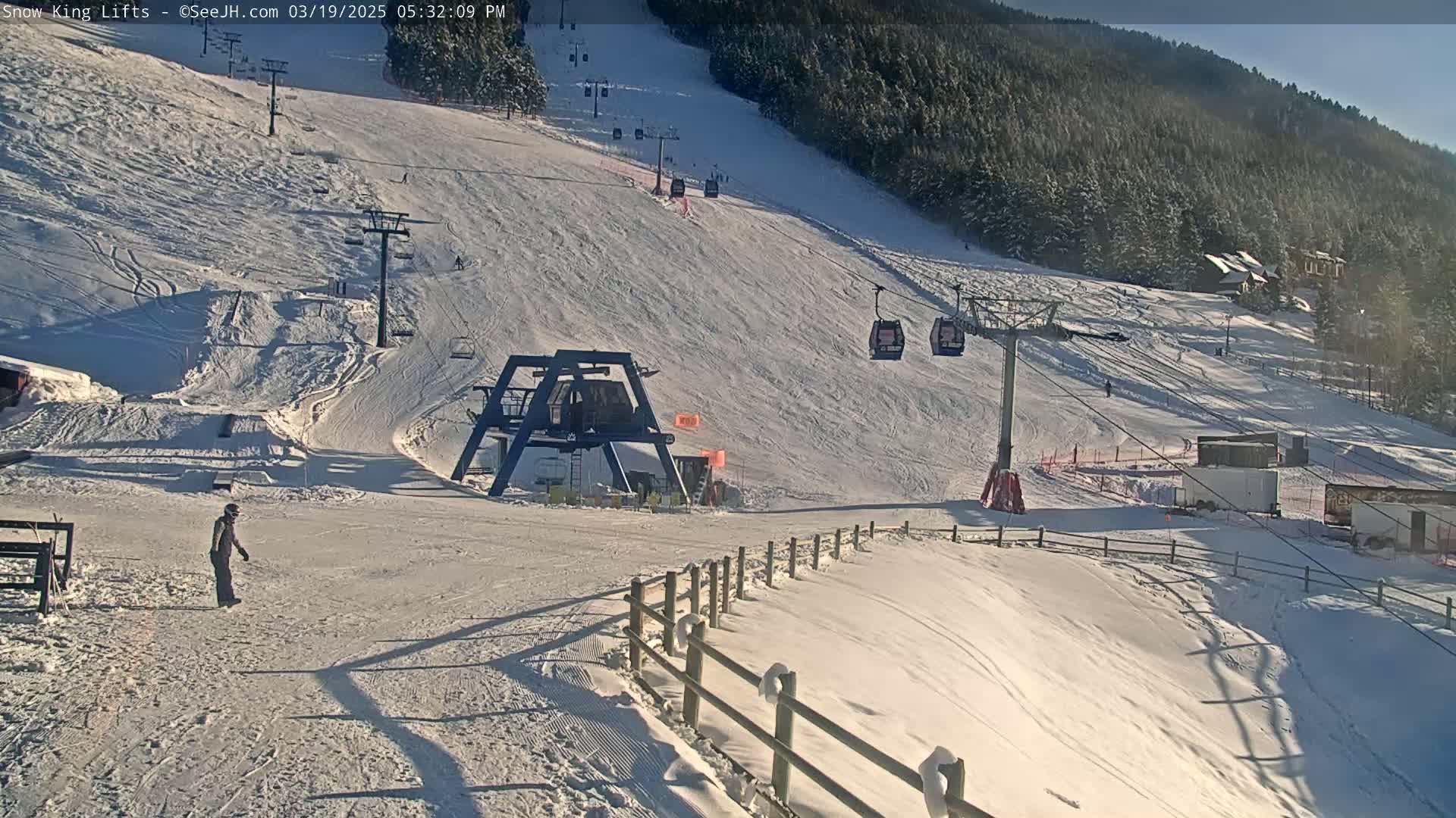 A snowy ski slope with several chairlifts, a few skiers, and a wooded mountain in the background under sunny conditions.