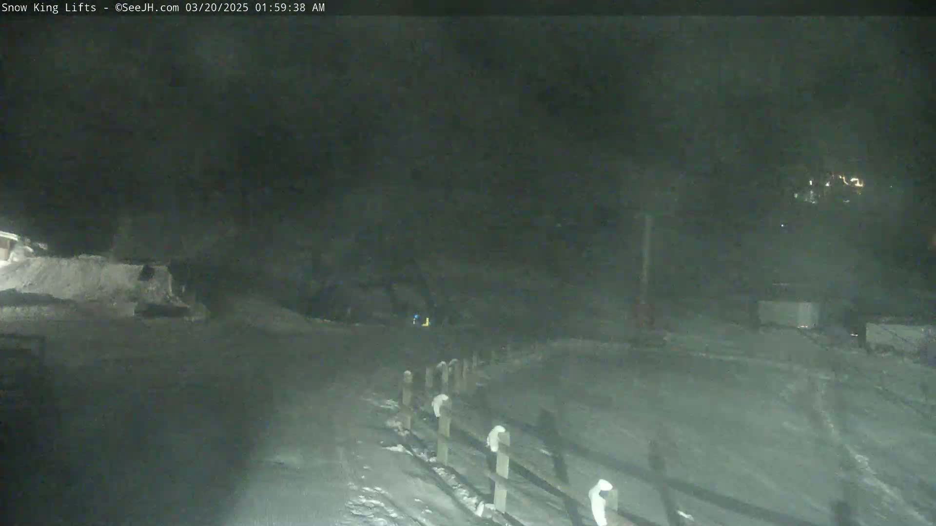 A snow-covered area at night, with a wooden fence and faint lights in the distance, under dark, possibly snowy conditions.