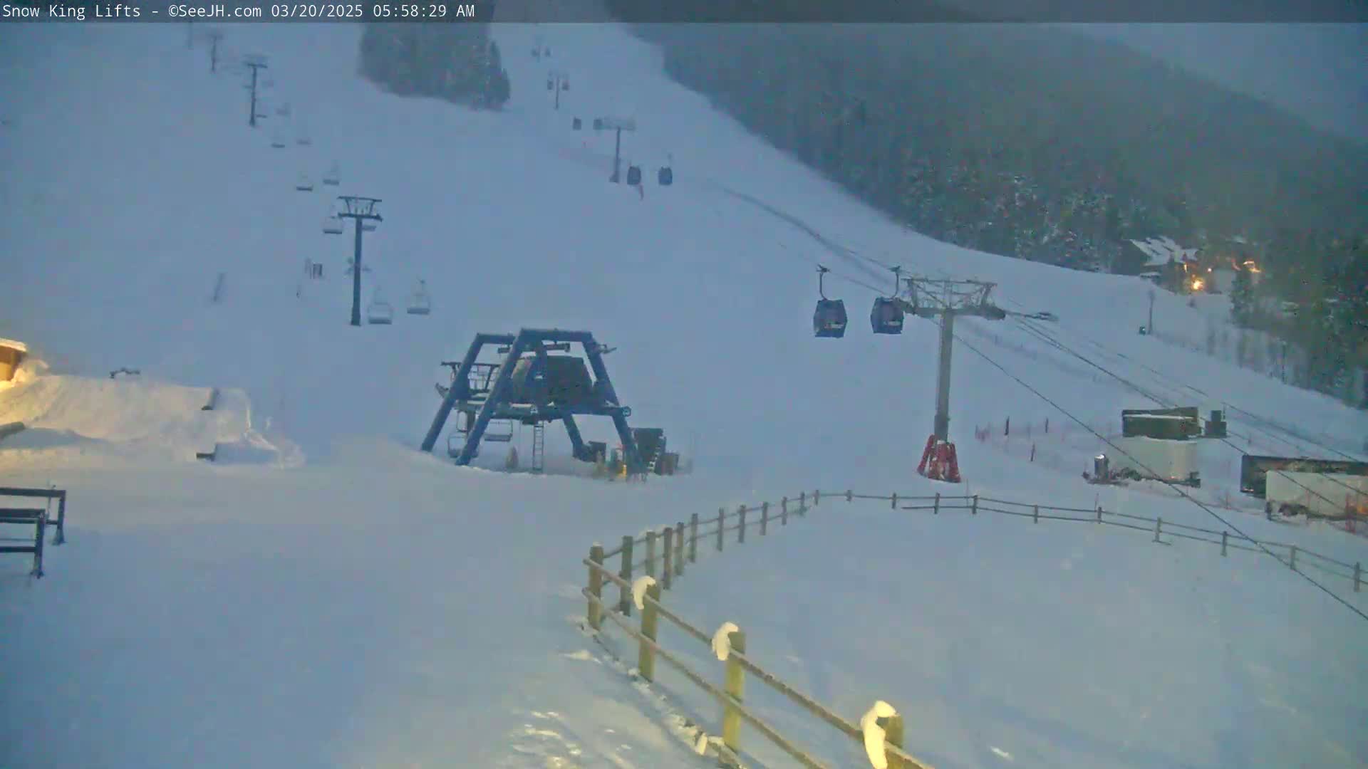 A snow-covered ski slope with chairlifts and a wooden fence under overcast skies.