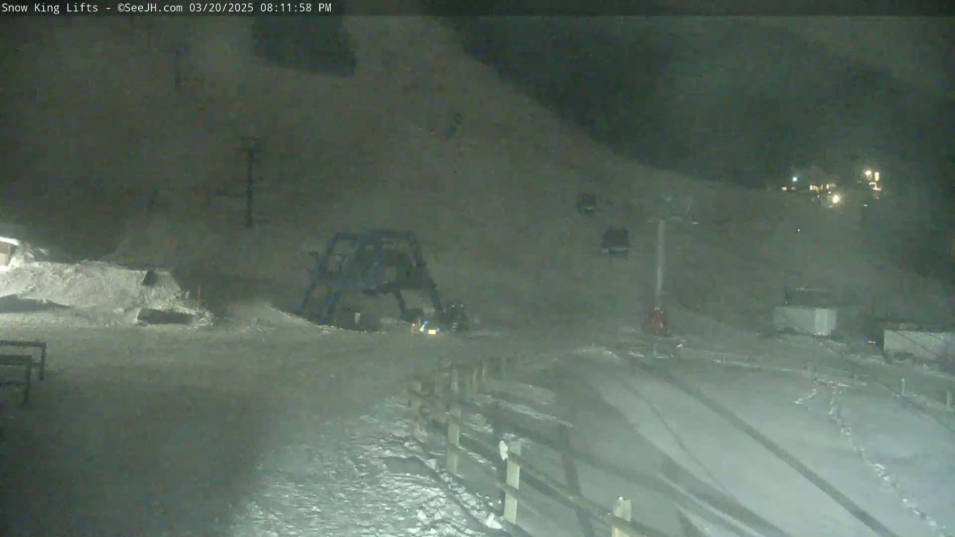 A nighttime, snow-covered ski slope scene shows a snowcat near a ski lift, with several other vehicles parked nearby under dim lighting.