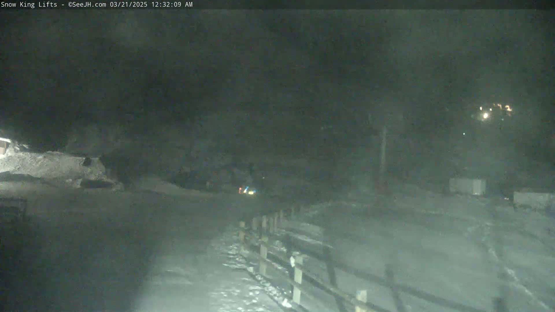 A nighttime snowy scene shows a road with a wooden fence, a few lights in the distance, and a vehicle partially visible in the middle ground.
