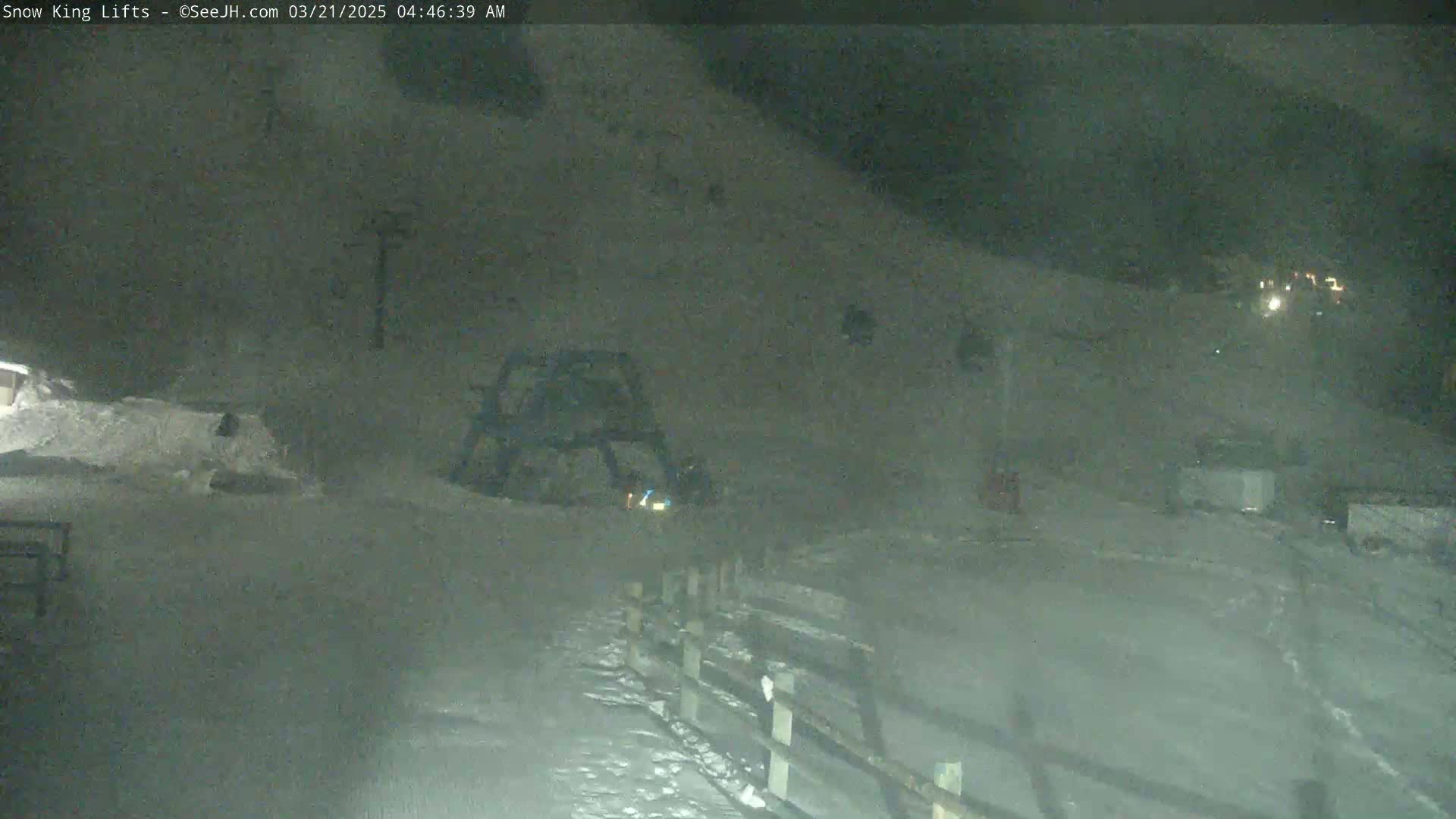 A snow-covered nighttime scene shows a ski lift and a snowcat near a wooden fence, with faint lights visible in the distance.