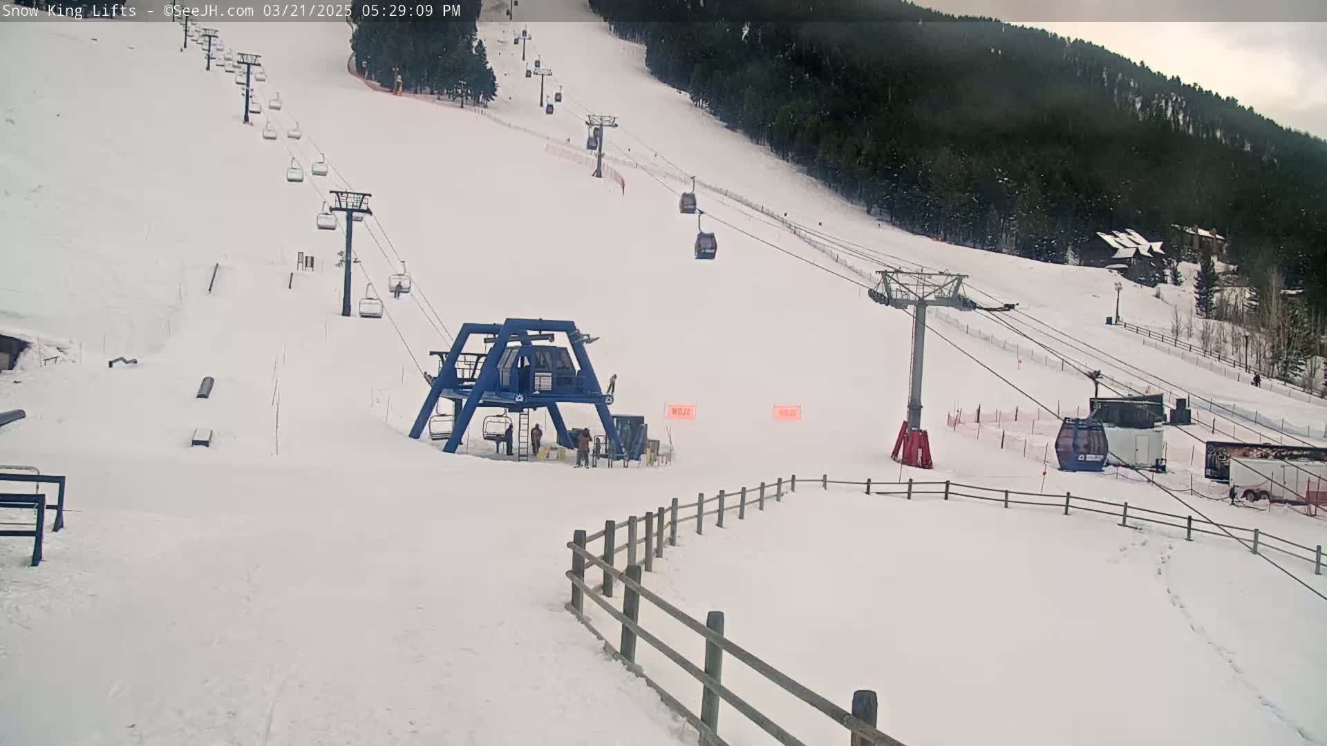 A snow-covered ski slope with several chairlifts and a few people near the base, under a cloudy sky with light snowfall.