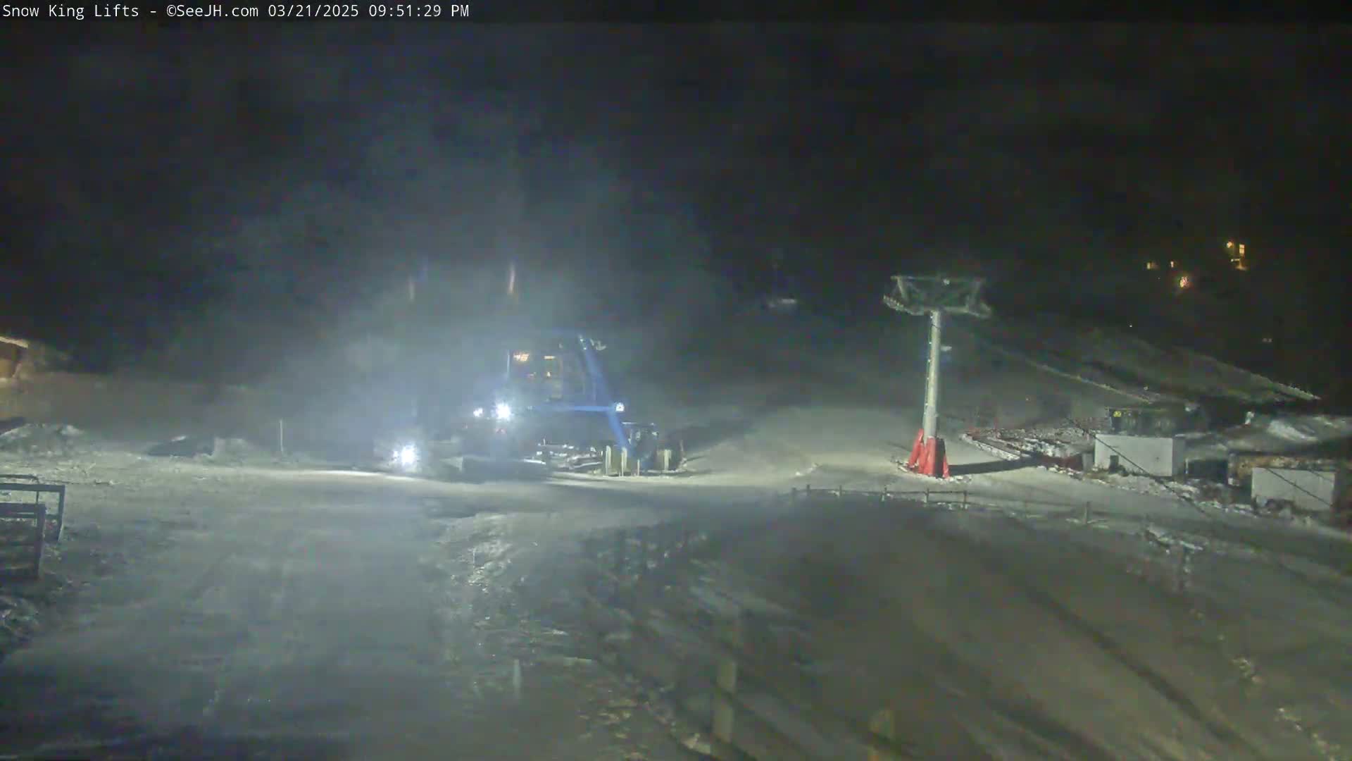 A snow groomer is working on a snow-covered slope at night, near a ski lift.