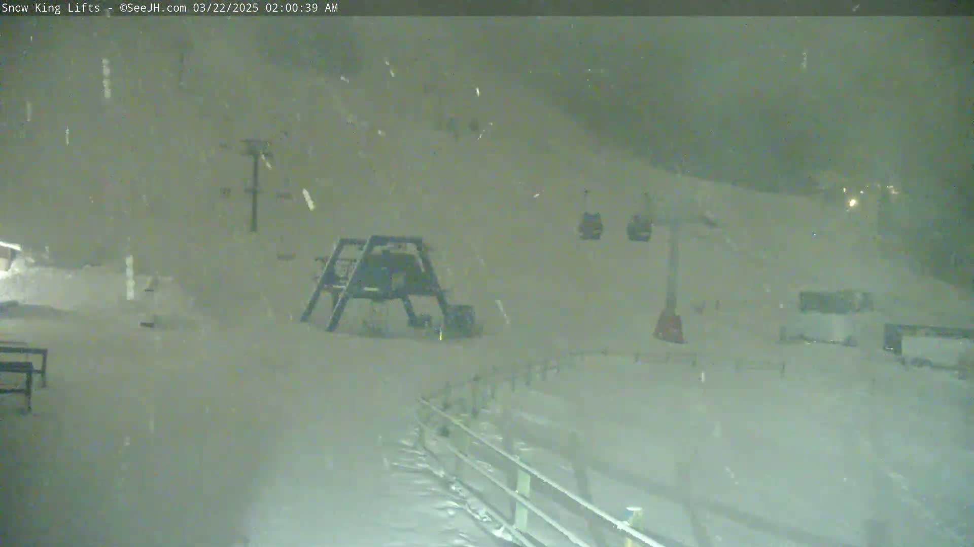 A nighttime snow scene shows a ski lift and several chairlifts covered in falling snow.